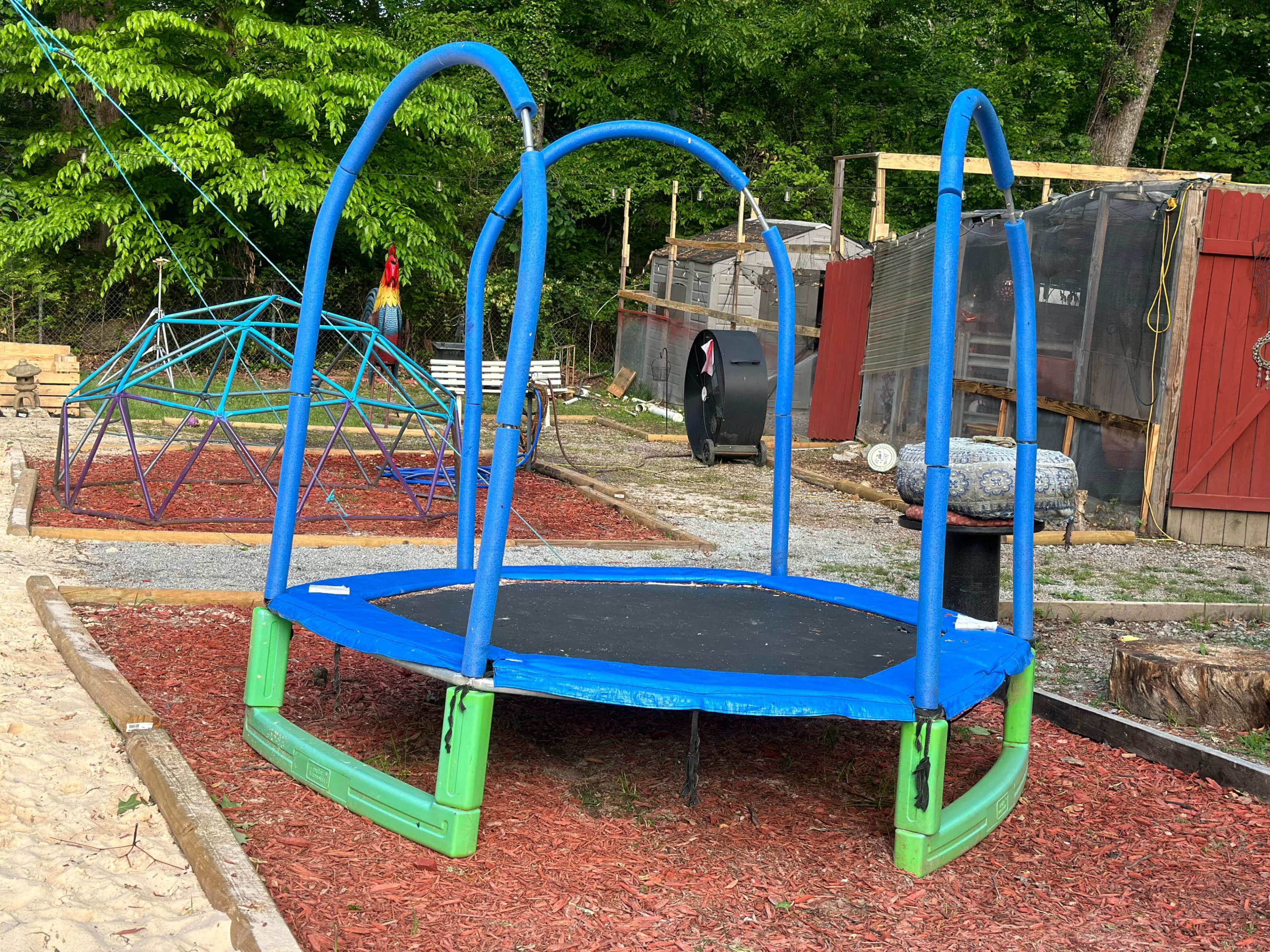 A blue and green trampoline is set in a playground area surrounded by mulch and playground equipment.