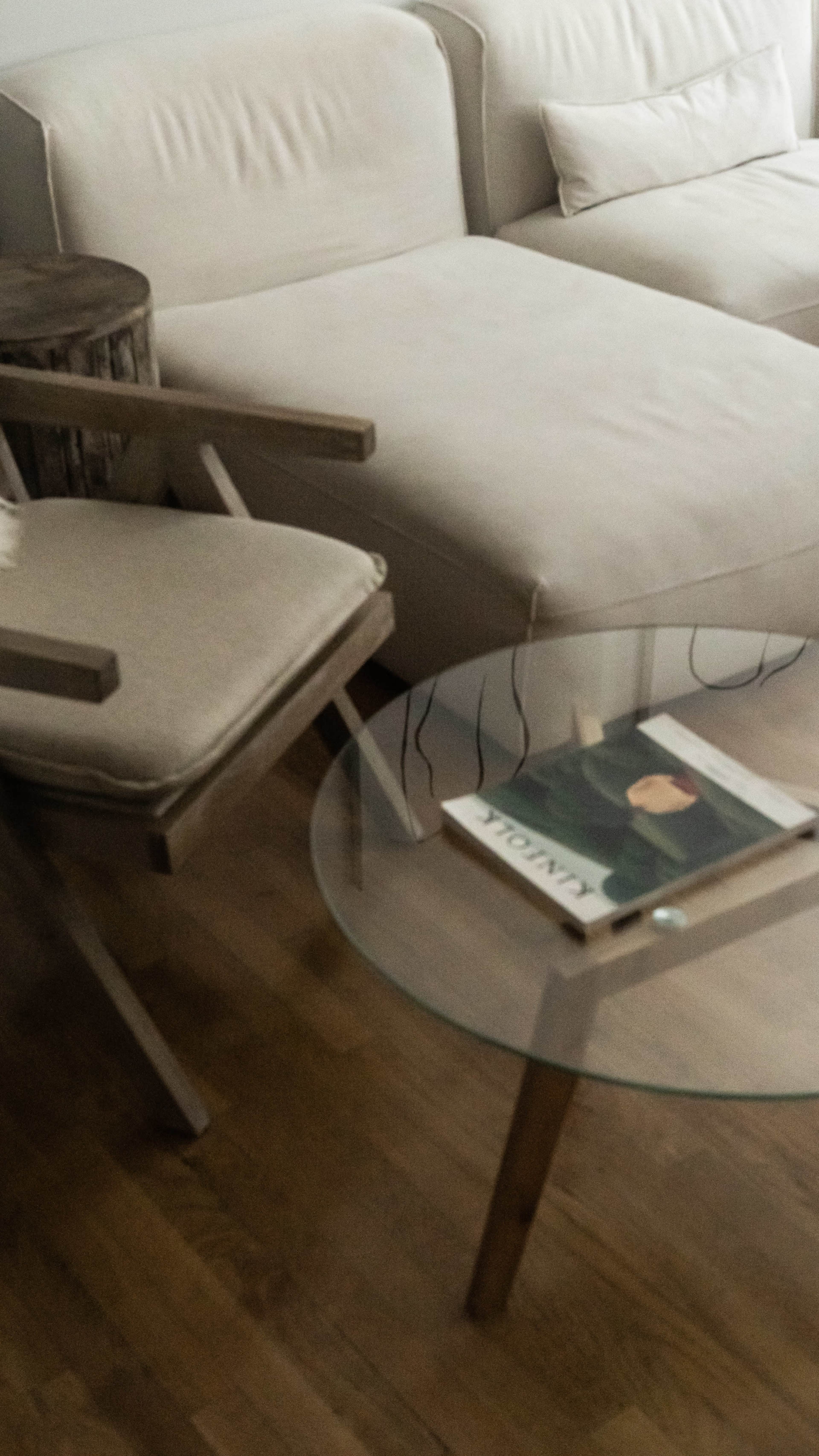 A wooden chair and a glass coffee table with a magazine on it are positioned next to a light-colored sectional sofa in a living room.