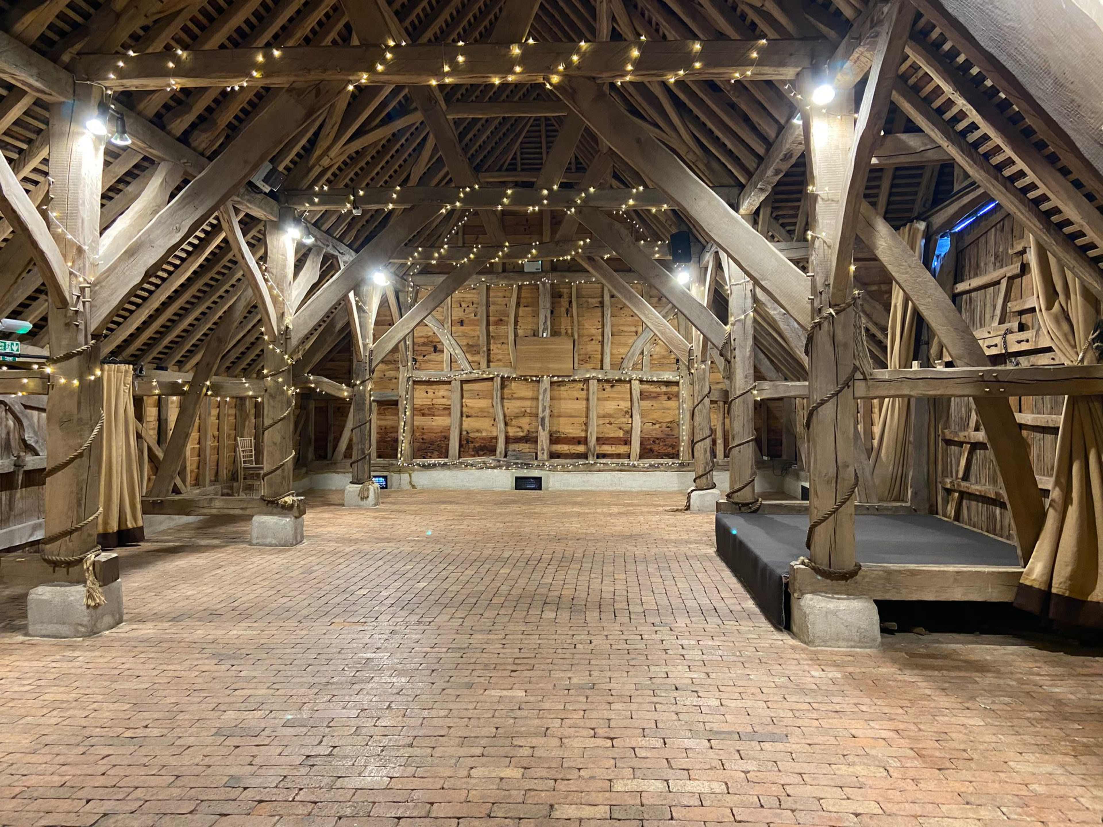 An empty barn interior with wooden beams, brick flooring, and string lights hanging from the ceiling.