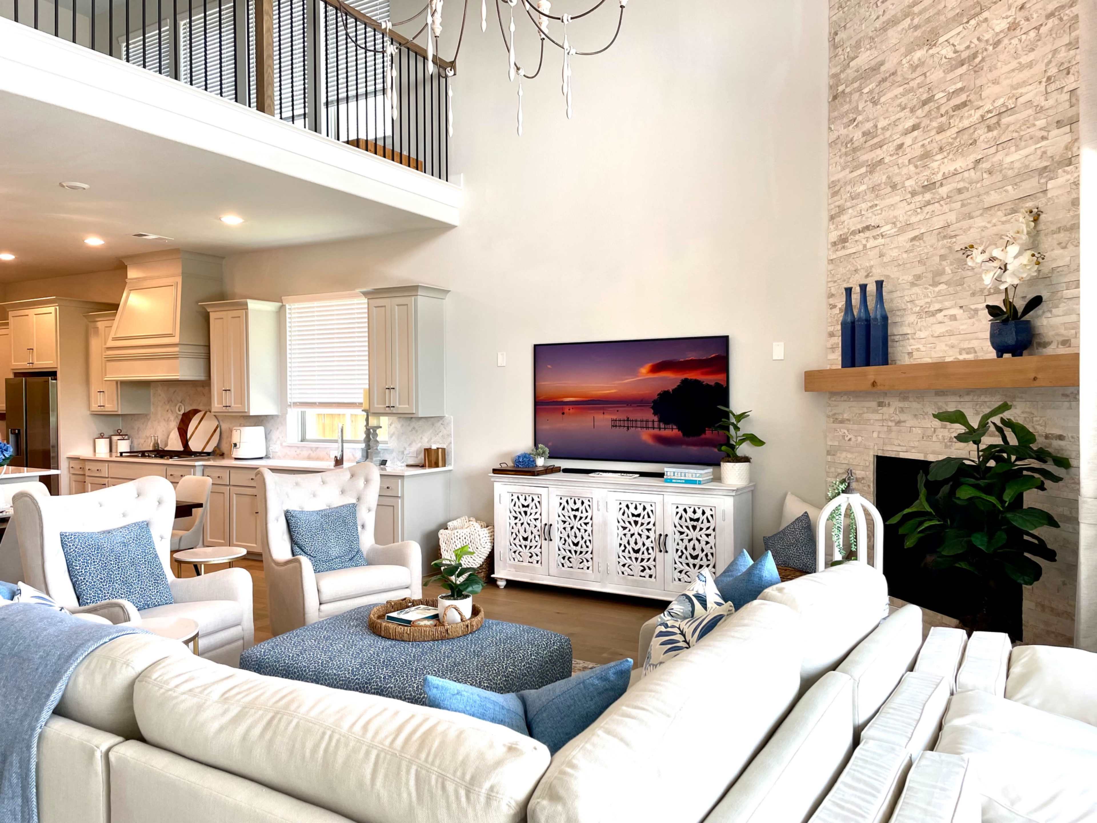 A modern living room features a sectional sofa, a decorative coffee table, and a wall-mounted TV above a white sideboard, with an adjacent kitchen visible in the background.