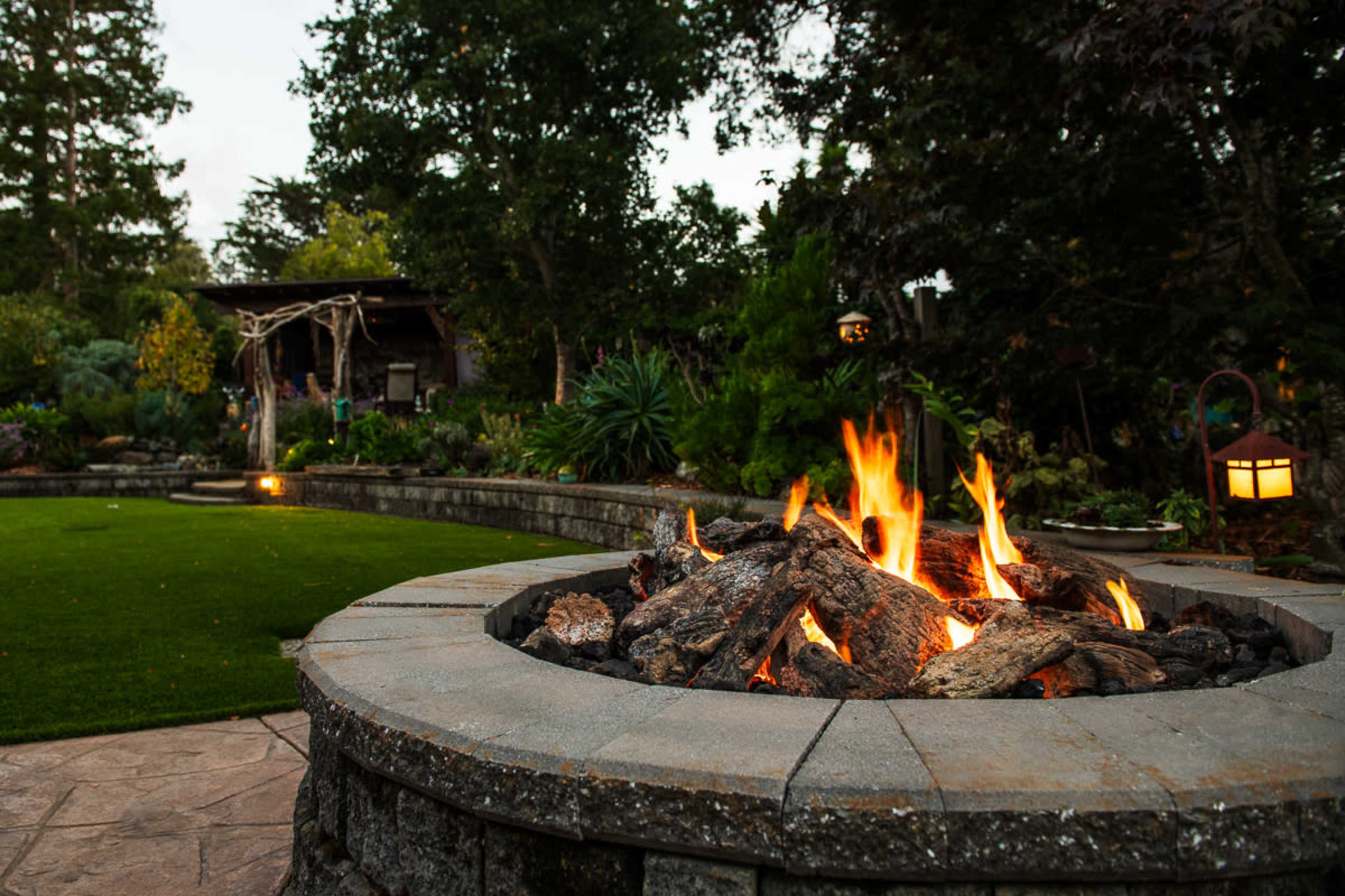 A circular stone fire pit with flames is surrounded by a lush garden and patio area.