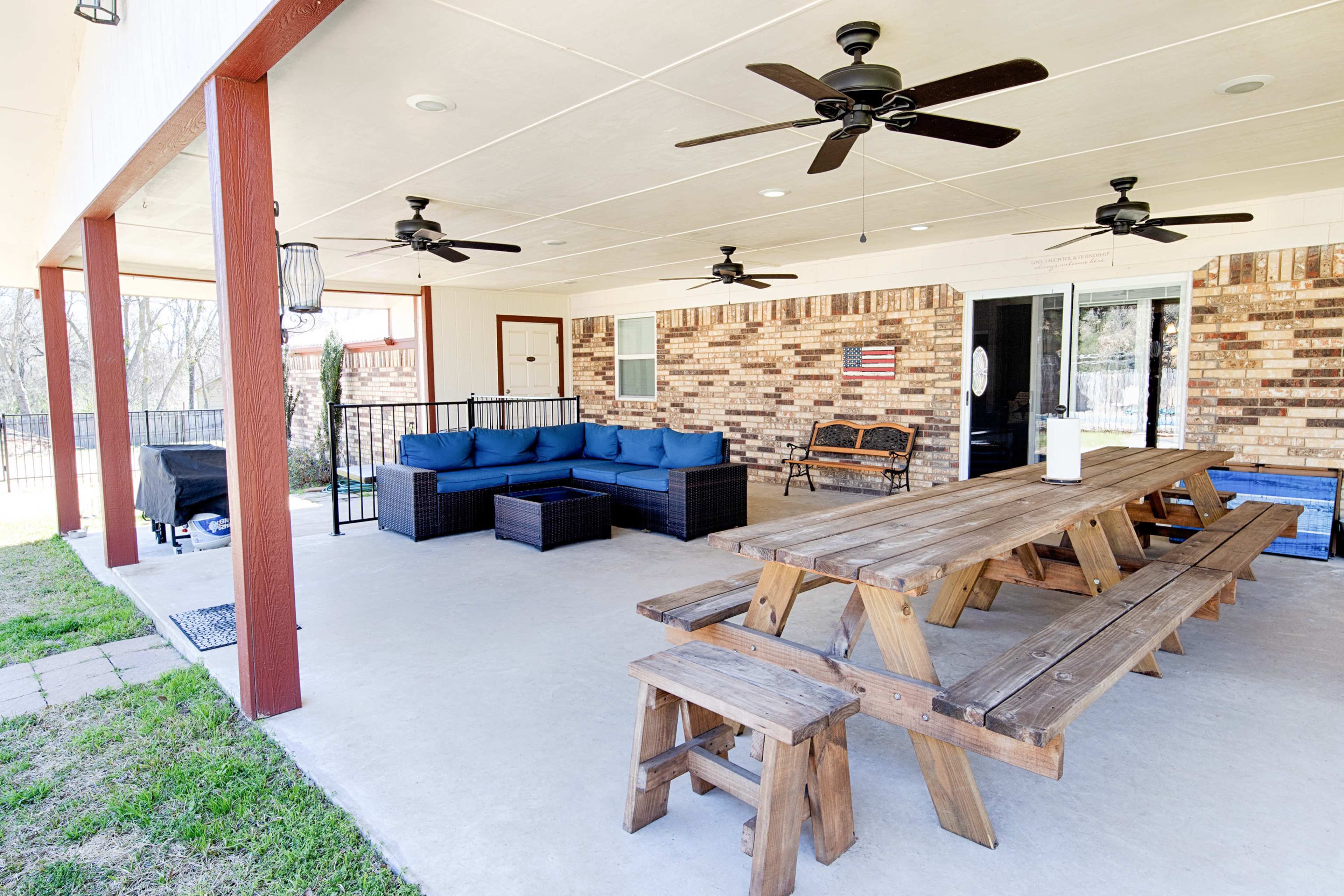 A covered outdoor patio featuring a large wooden dining table, a seating area with blue cushions, and multiple ceiling fans.