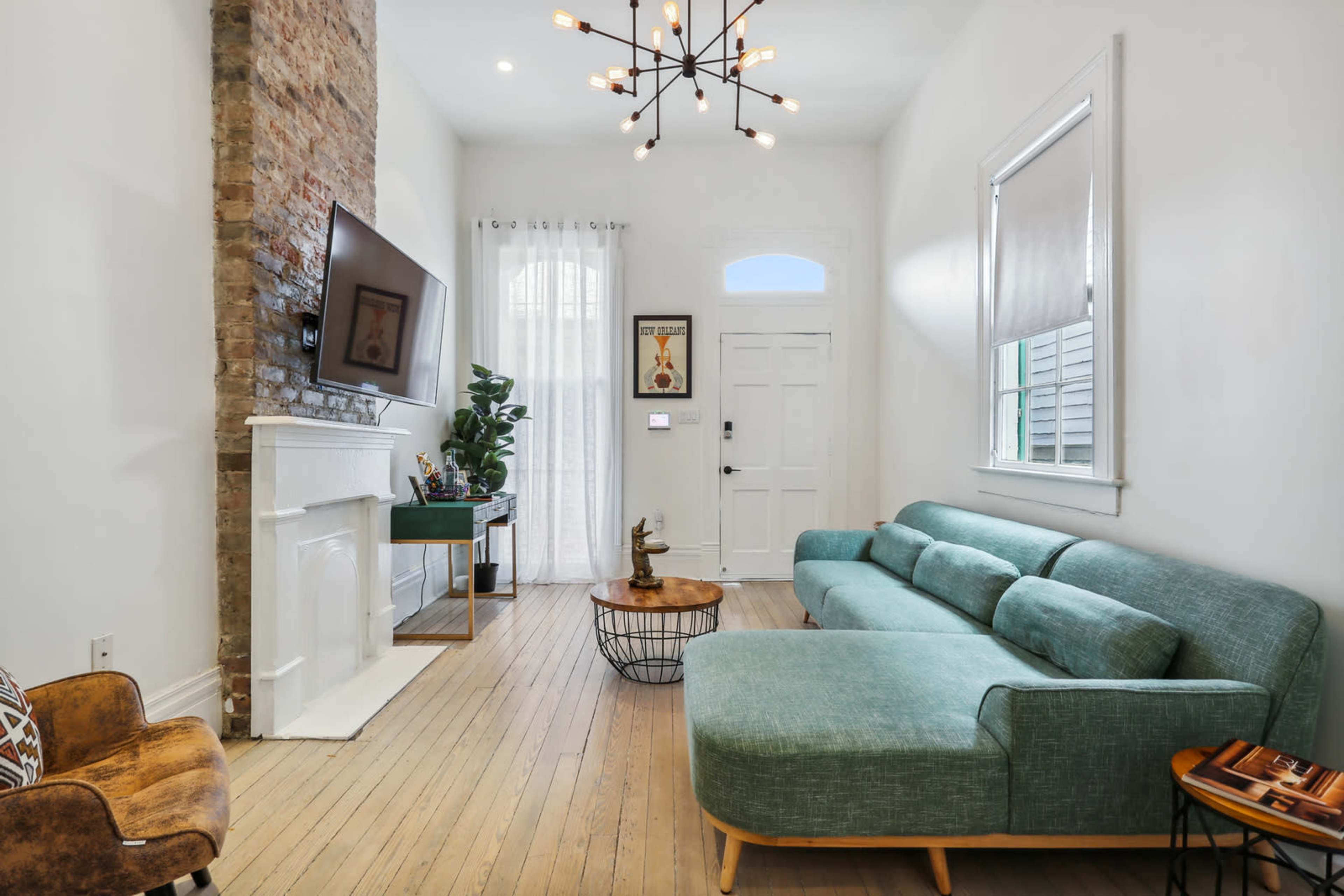 A modern living room features a green sectional sofa, a wood coffee table, a mounted television, and a large window with sheer curtains.