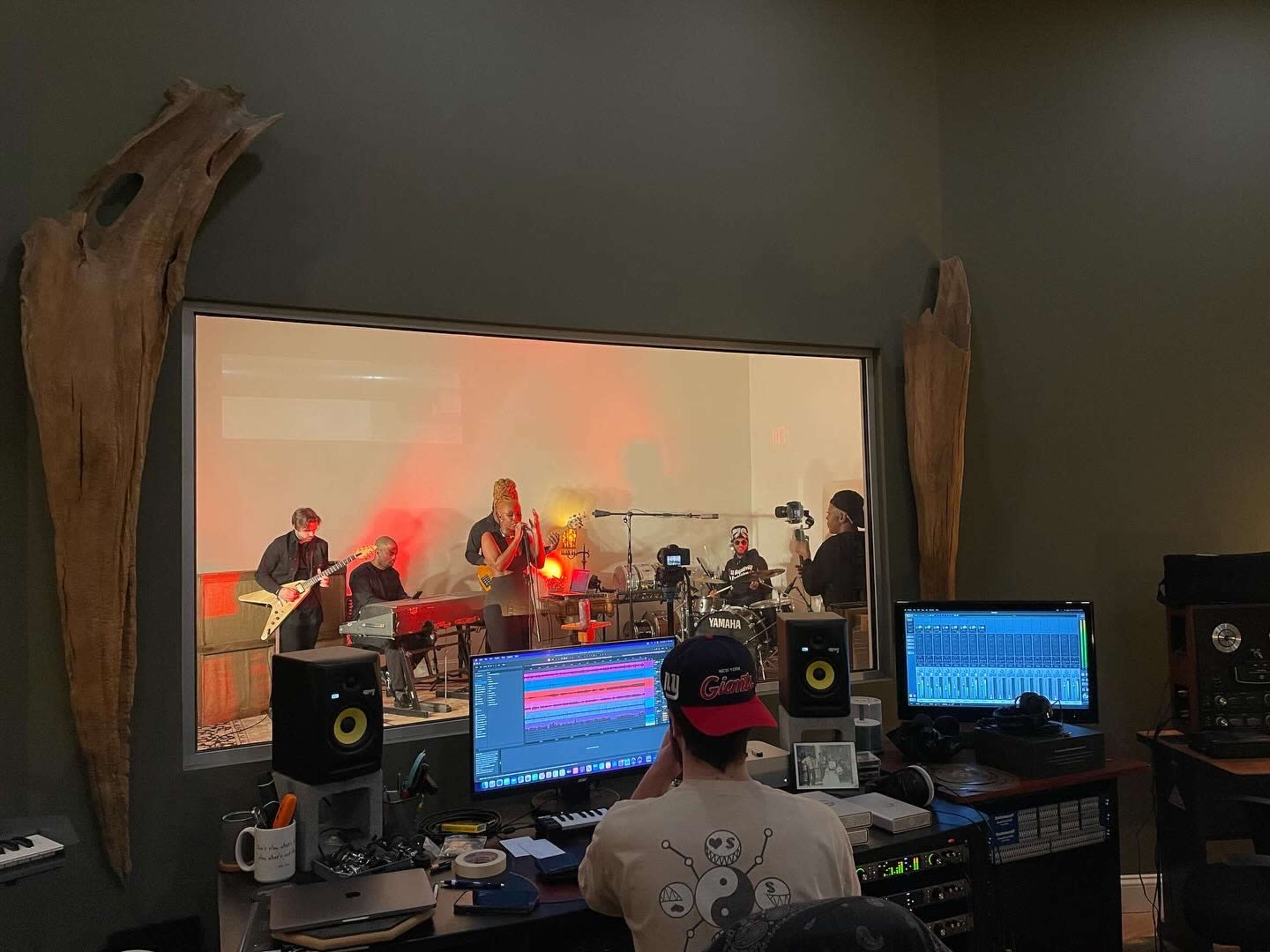 A band performs in a studio behind a glass wall, while a person works on a music production setup in the foreground.