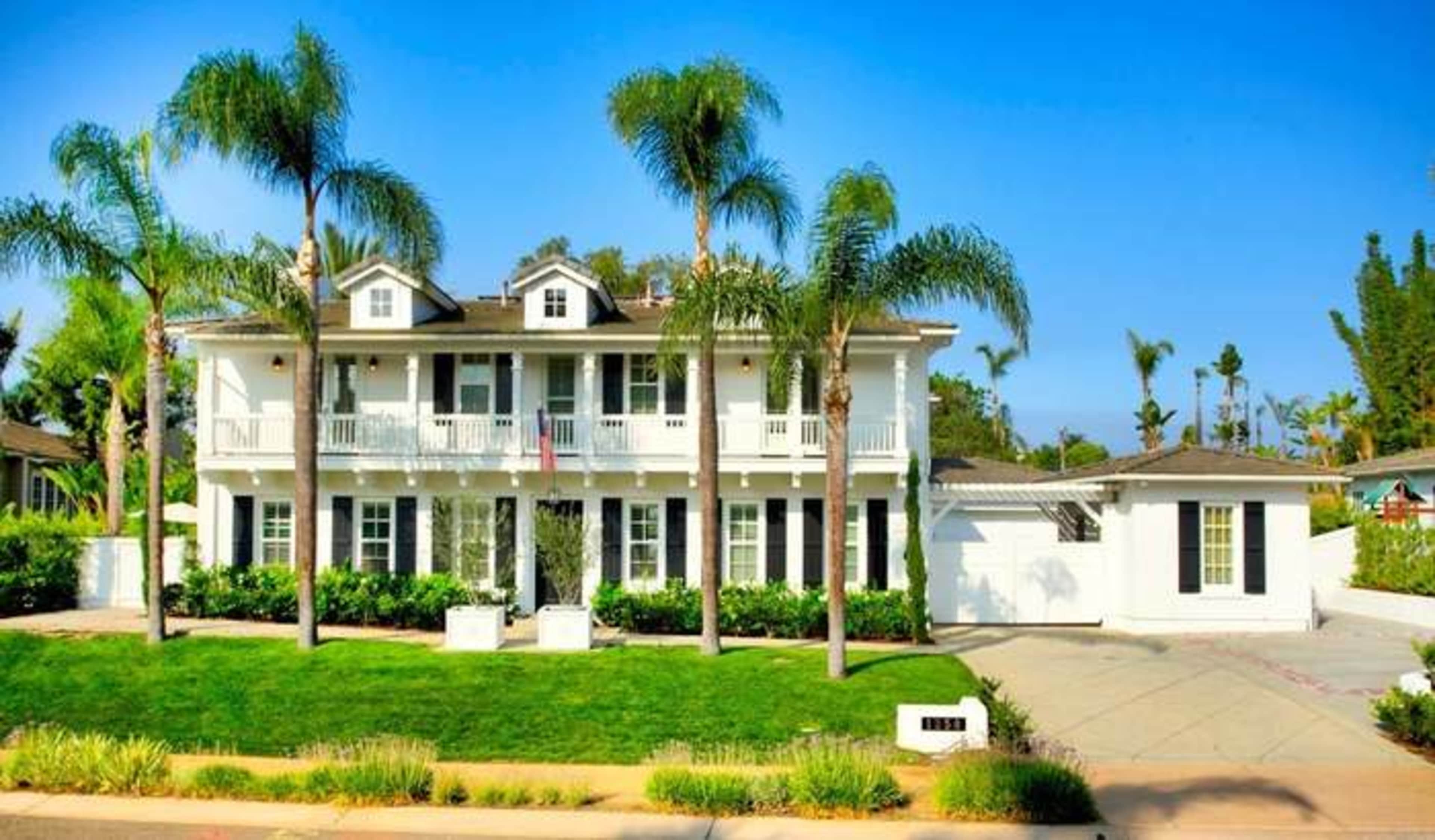 A two-story white house with a front porch and palm trees in the yard.
