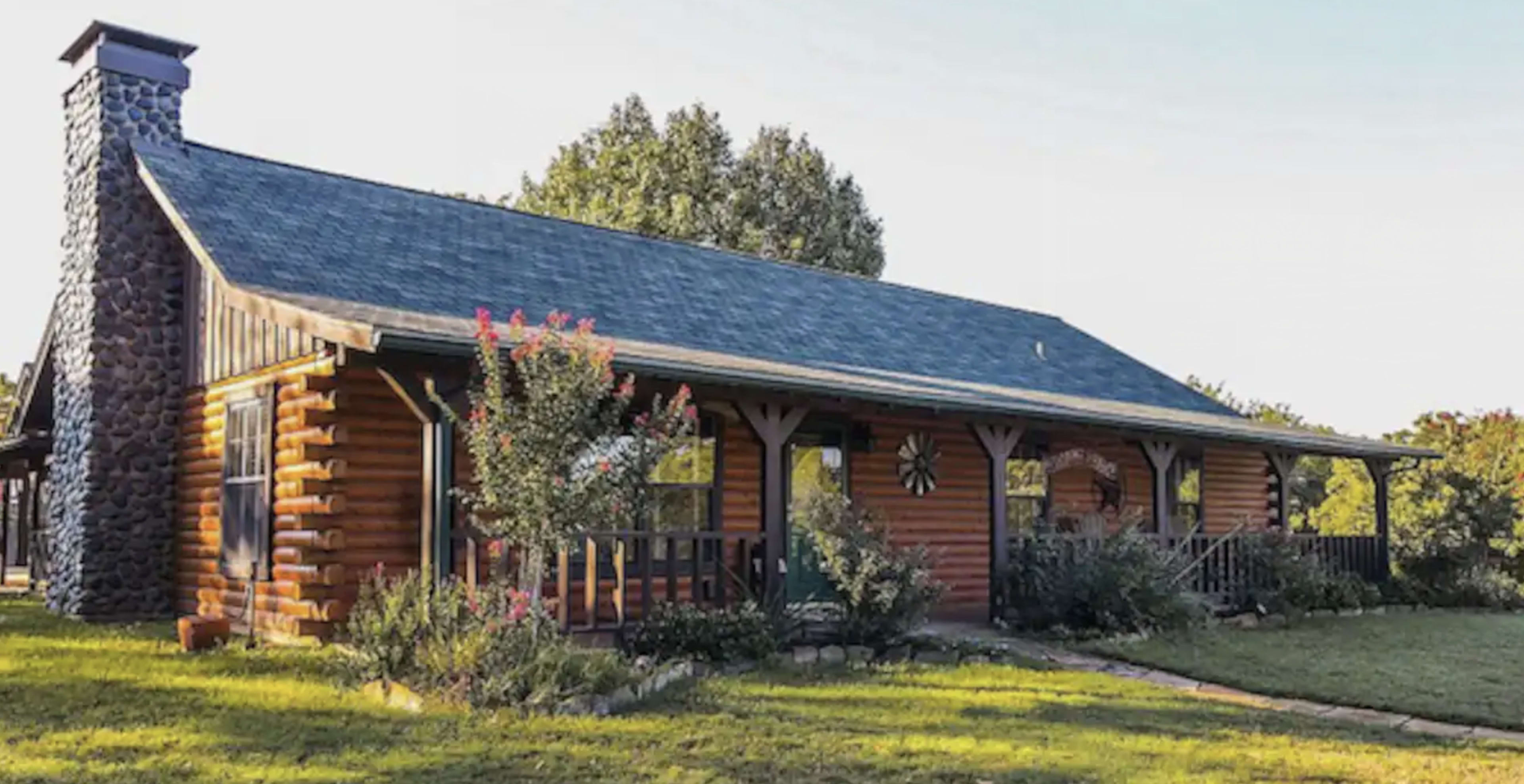 A log cabin with a stone chimney and a porch is surrounded by greenery and a well-maintained lawn.