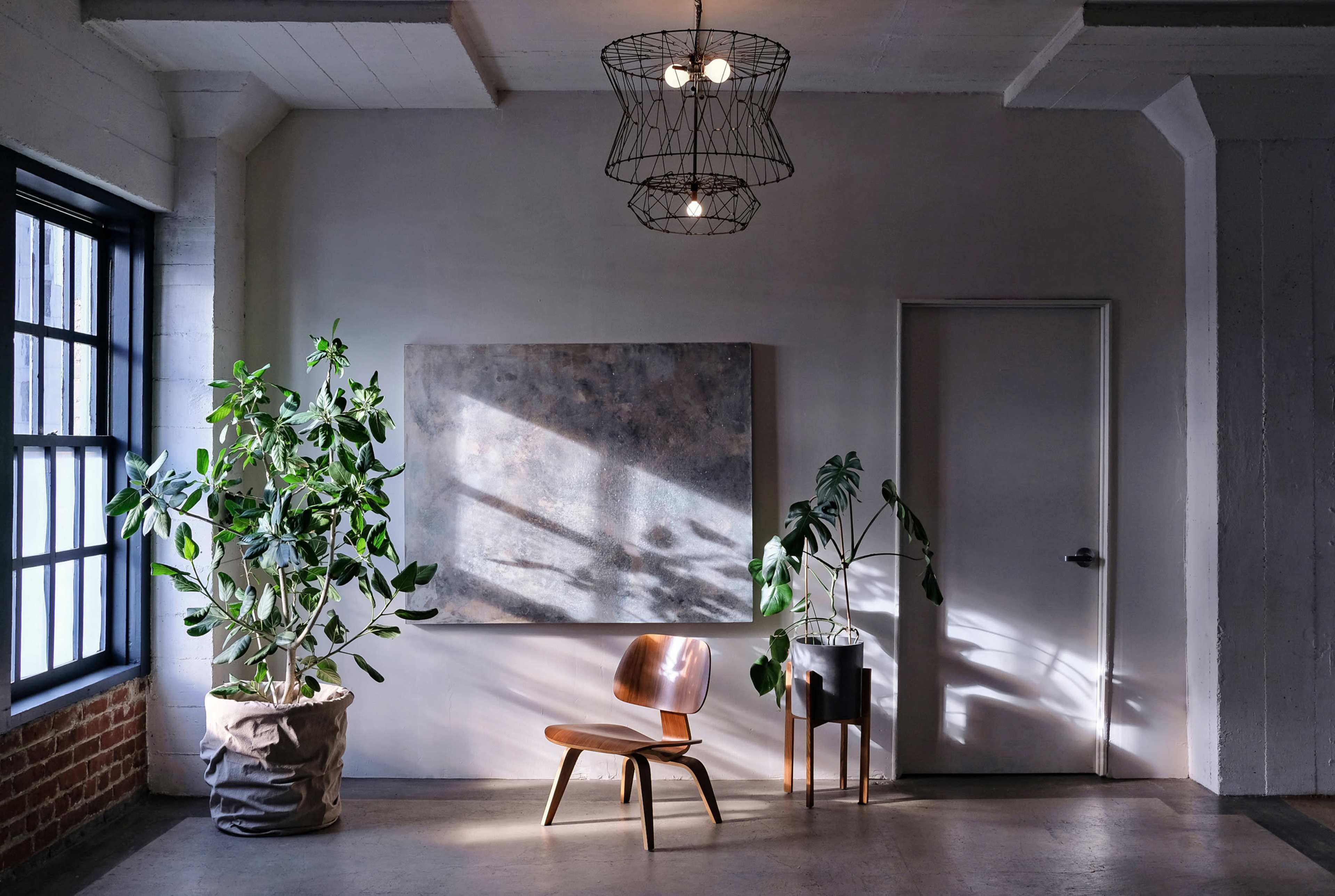 A wooden chair is positioned under a pendant light next to two potted plants and a large abstract painting on a wall in a well-lit room.