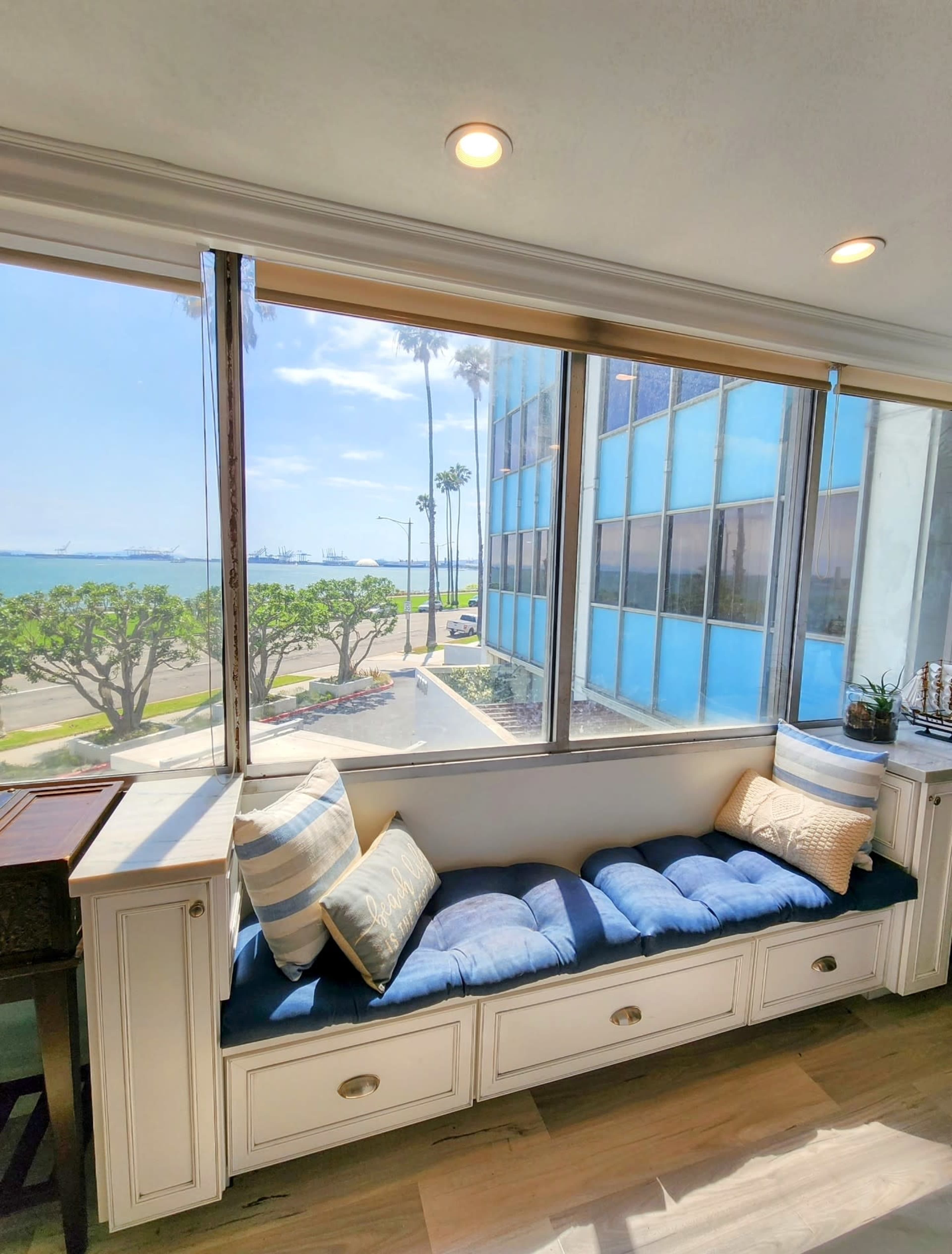 A bright room features a cushioned window seat with decorative pillows, offering a view of palm trees and a waterfront area outside.