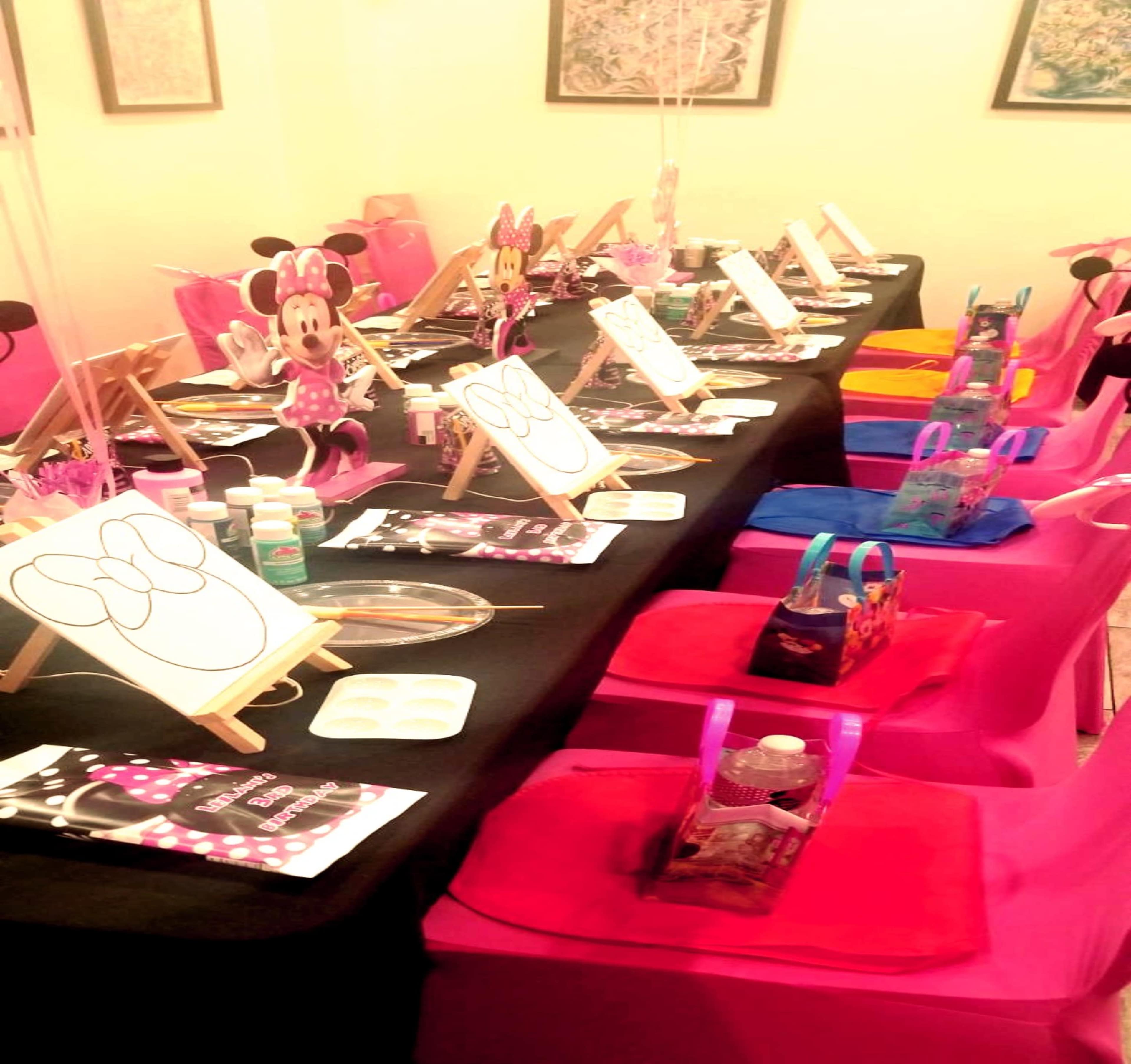 A colorful party setup features a long table adorned with art supplies and themed decorations, with matching chairs lined up on either side.