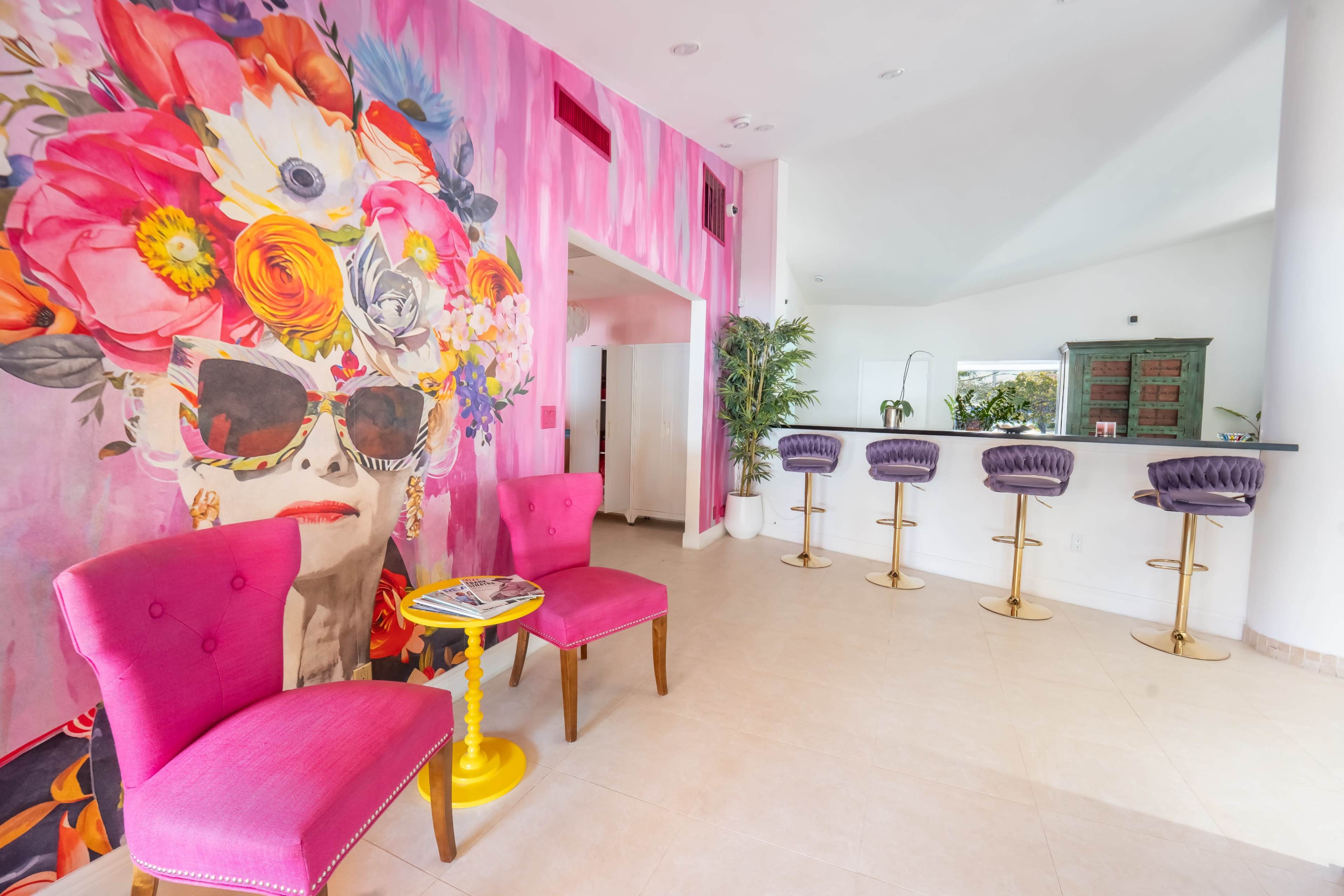 The image shows a vibrant interior space featuring a large floral mural, two pink chairs, a small yellow table, and a bar with purple stools.