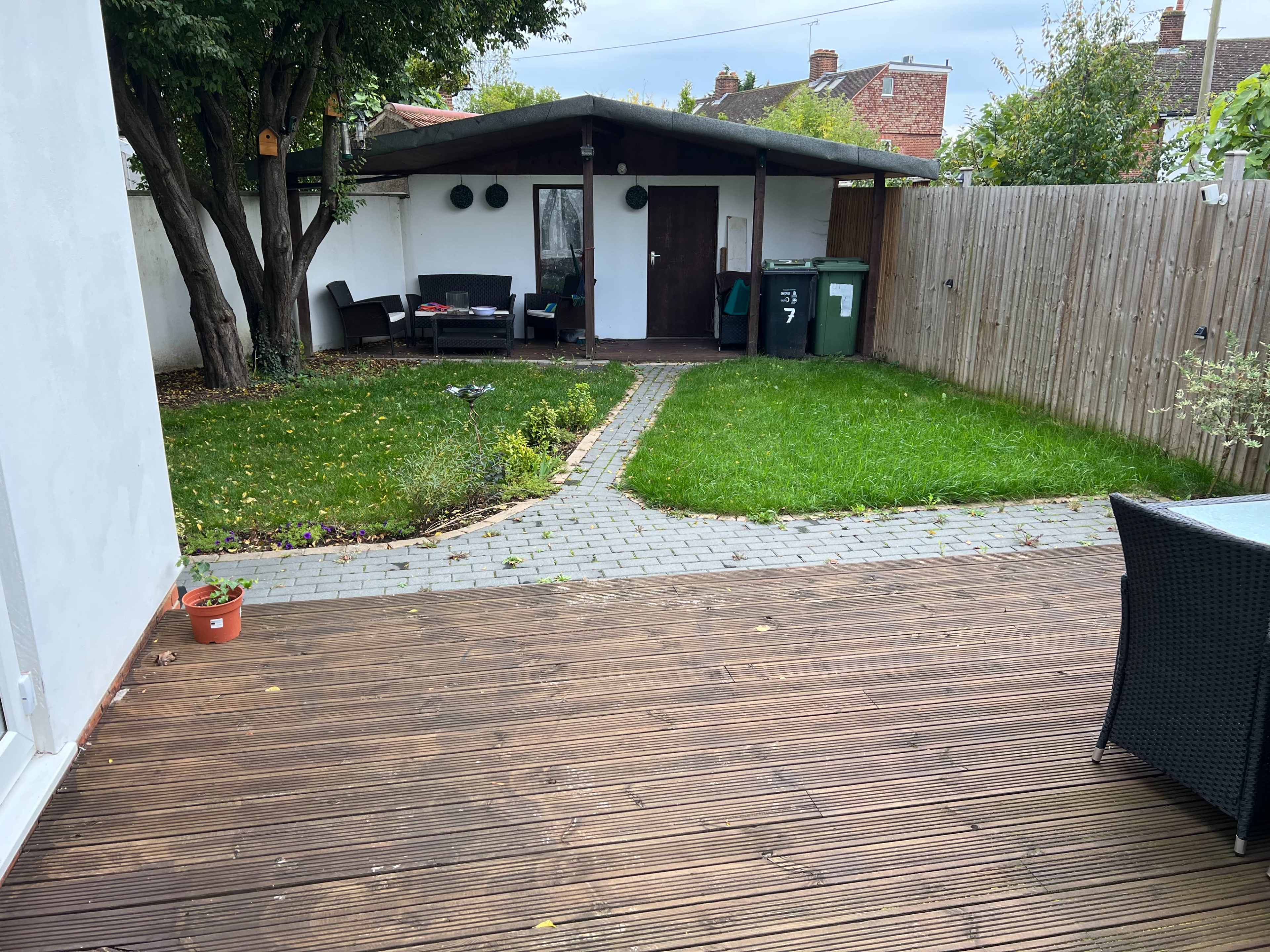 A backyard with a wooden deck, a stone path leading to a garden shed, and a grassy area surrounded by a wooden fence.