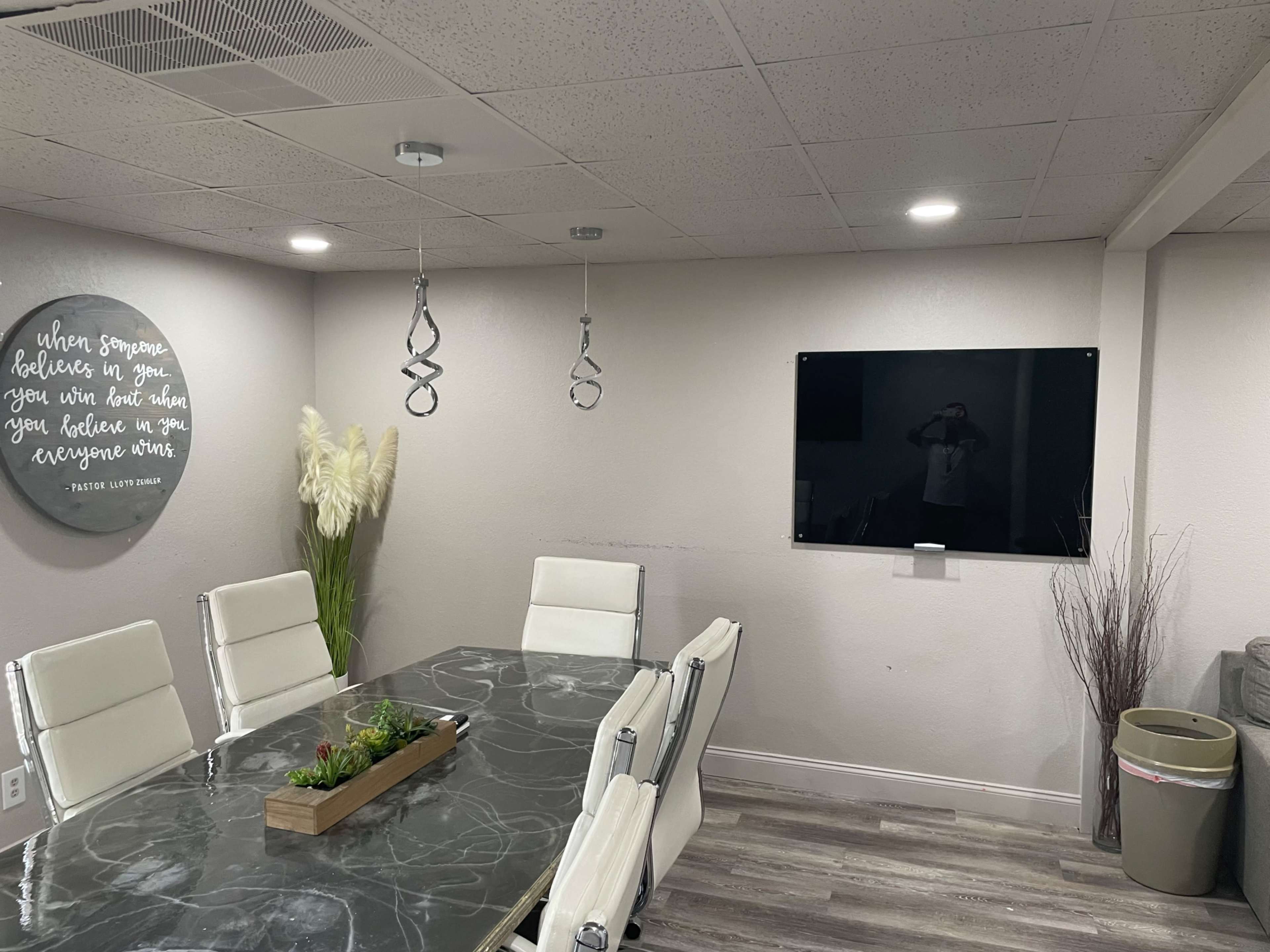 The image shows a modern conference room with a marble table, white chairs, a wall-mounted television, and decorative elements including plants and a circular wall art piece.