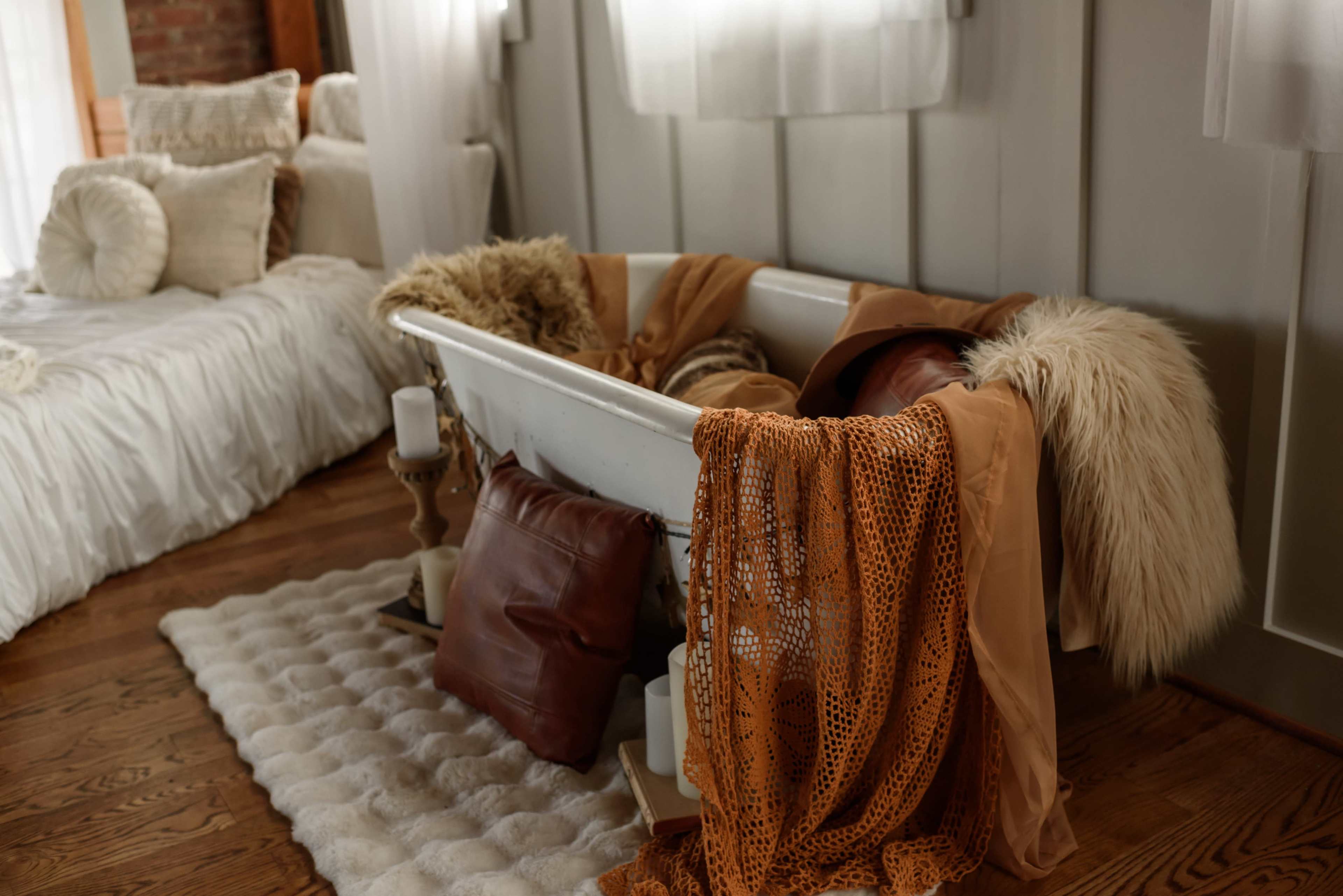 A vintage-style bathtub adorned with soft textiles and pillows sits on a wooden floor, surrounded by natural light filtering through sheer curtains.