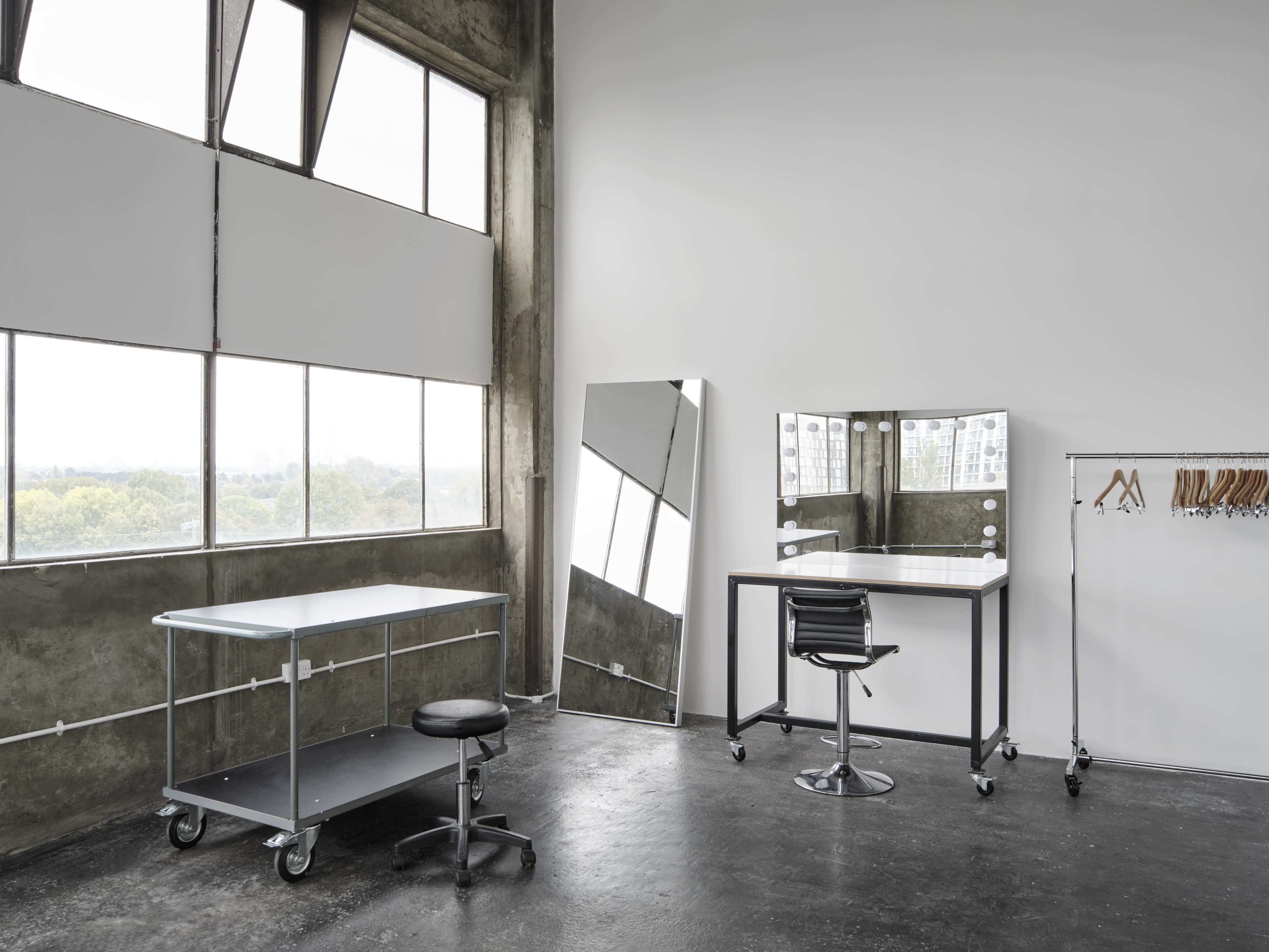 The image shows a modern, minimalistic workspace featuring a large mirror, a desk with a stool, and a clothing rack near large windows.
