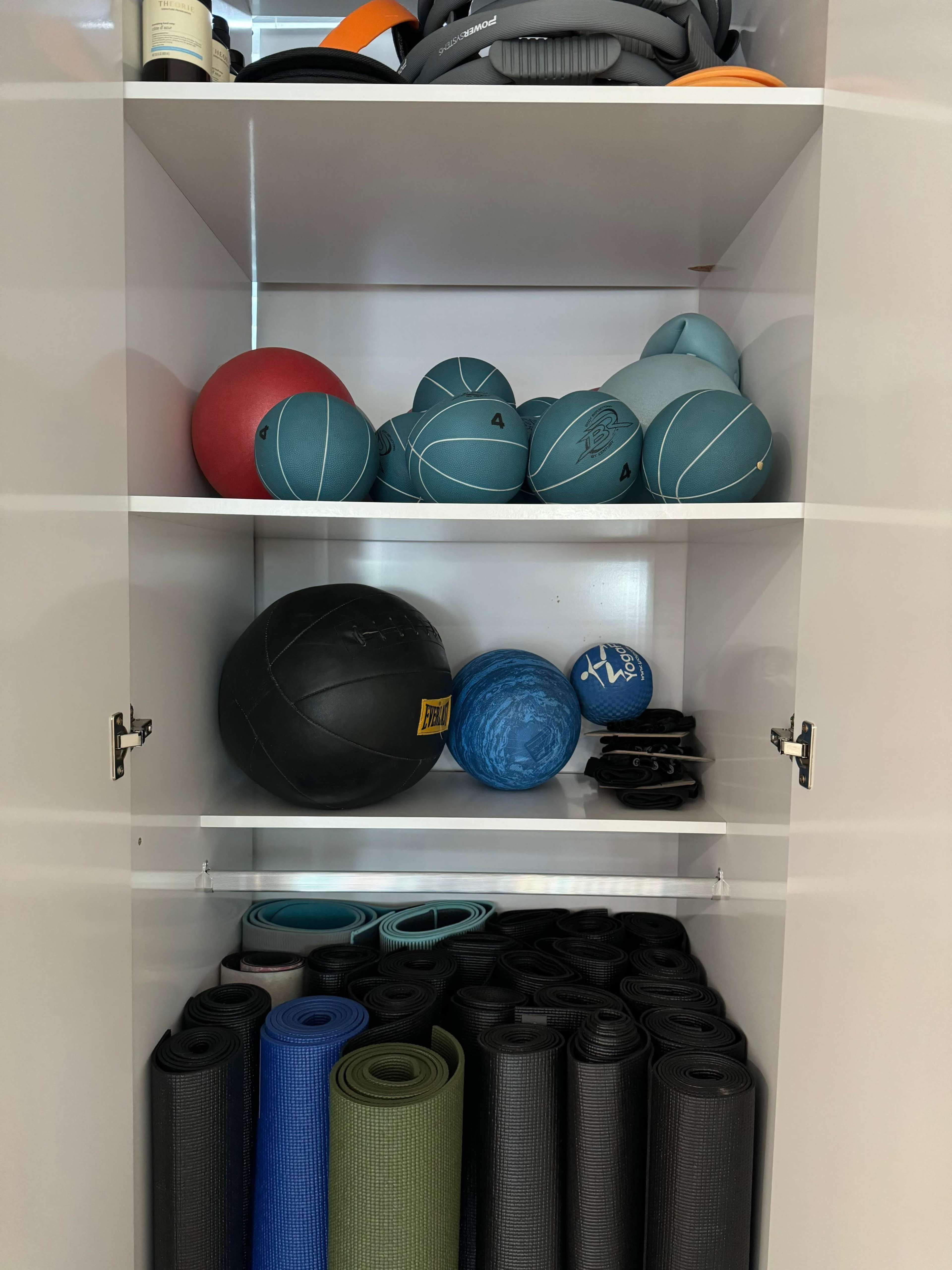 A white storage cabinet contains various fitness equipment, including medicine balls and exercise mats, organized on multiple shelves.