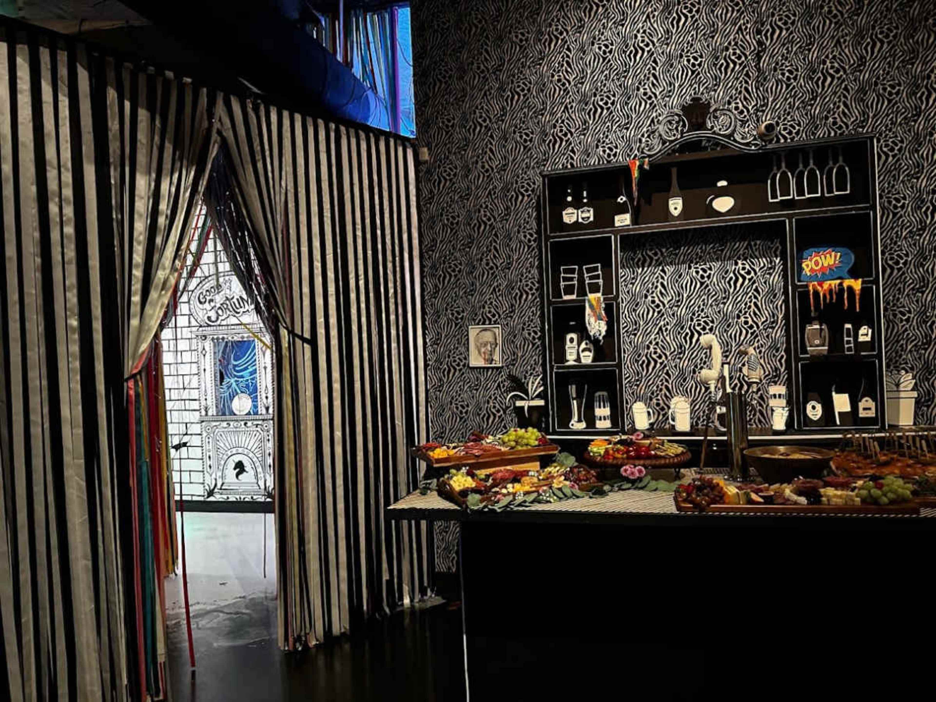 The image shows a room with a patterned wall, a table laden with various foods, and a backdrop featuring striped curtains and a decorative shelf.