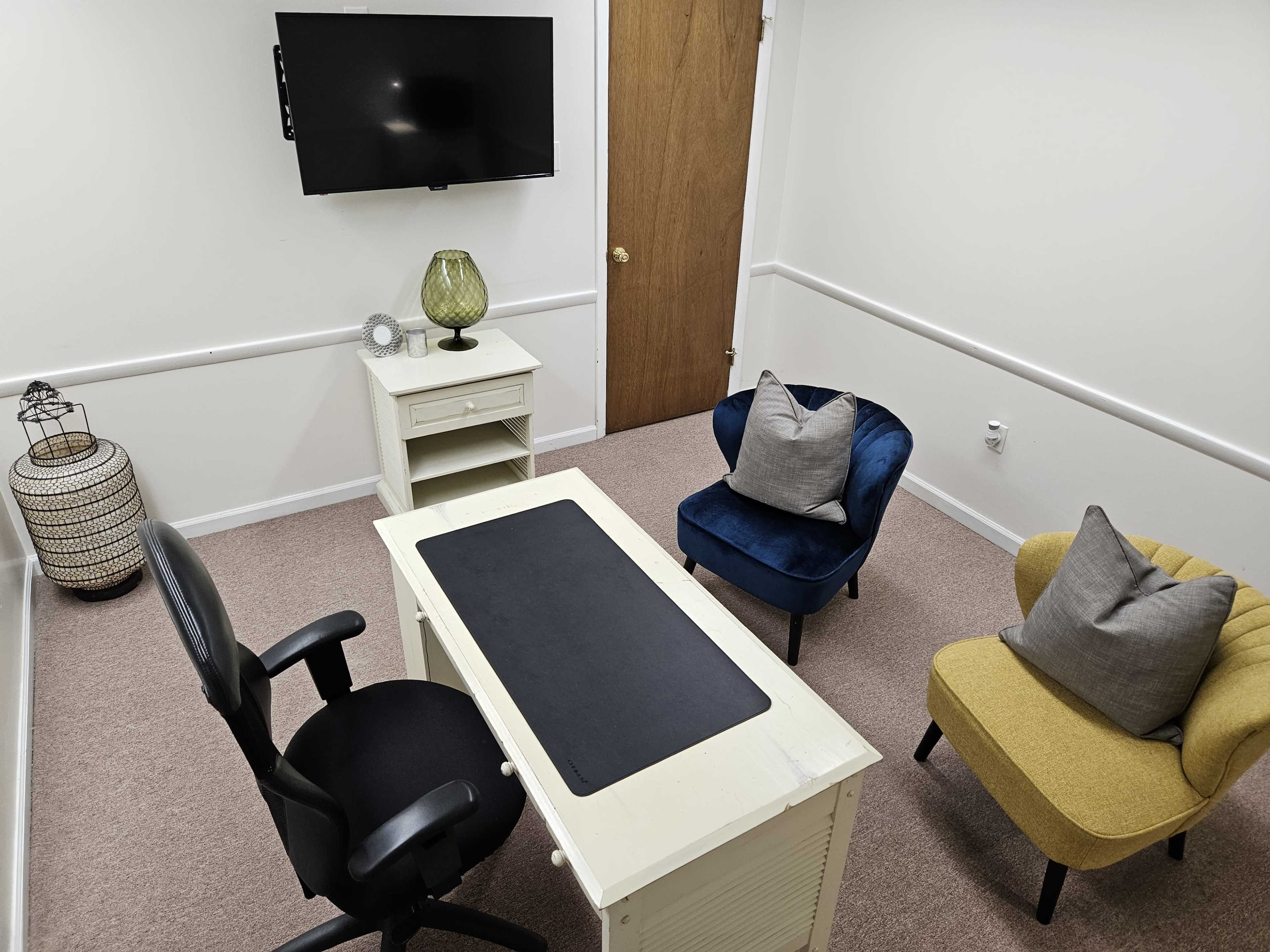 The image shows a small office space with a desk, two chairs, a TV mounted on the wall, and a decorative lamp.