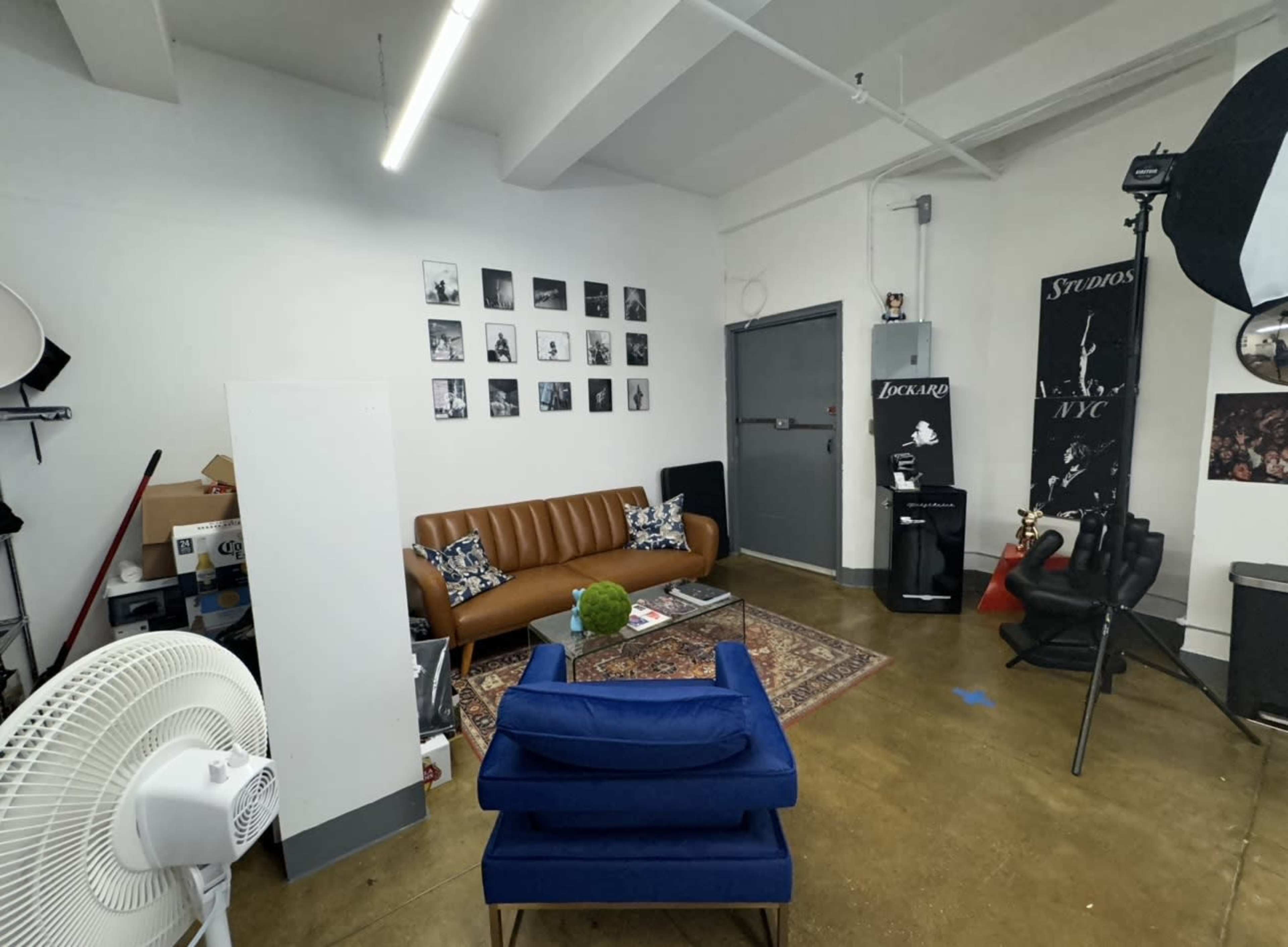 A small studio space featuring a brown leather sofa, a blue armchair, various framed black-and-white photographs on the wall, and a fan in the corner.
