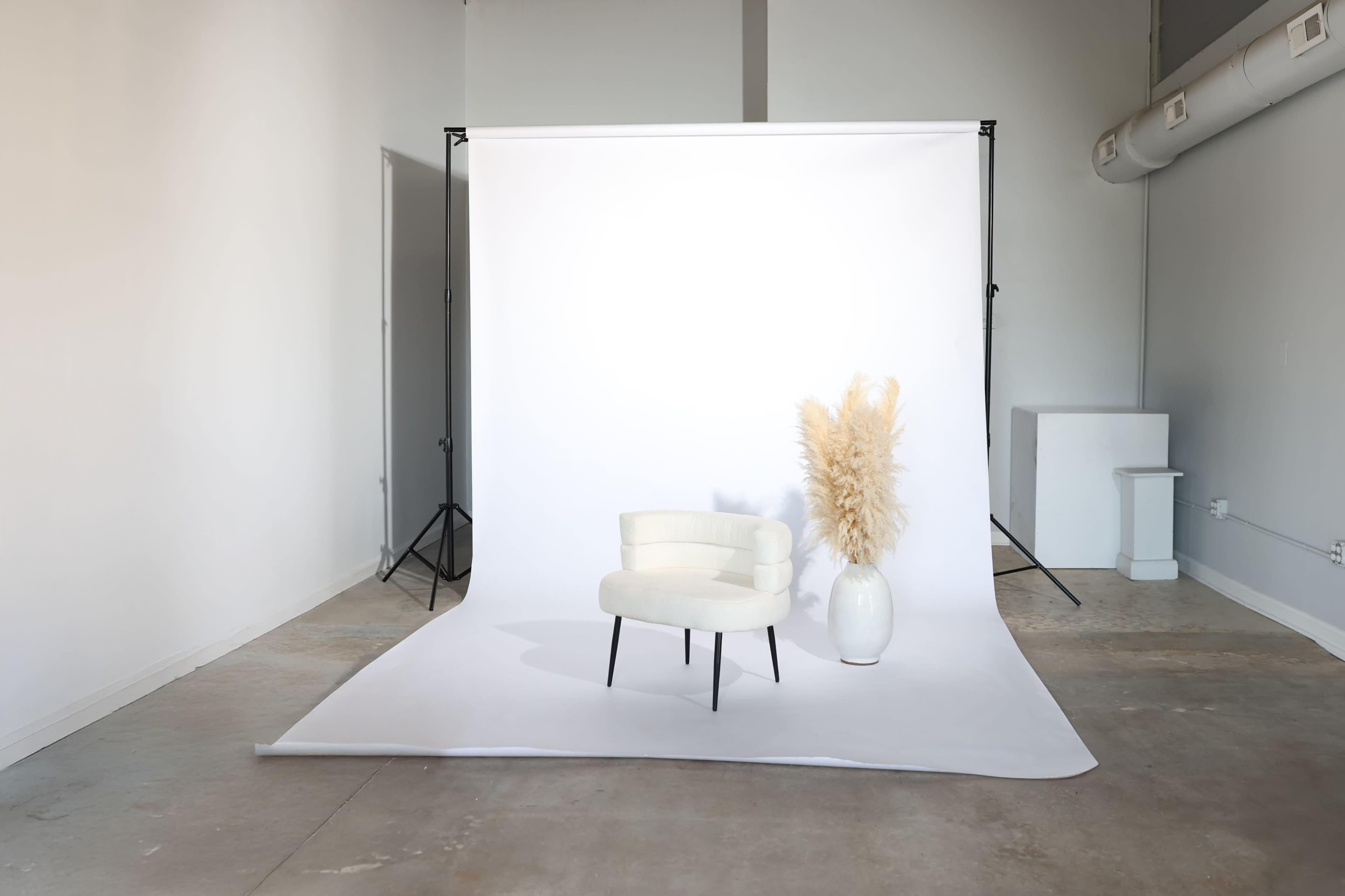 The image shows a minimalist studio setup featuring a white backdrop, a light-colored chair, and a tall vase with dried pampas grass.