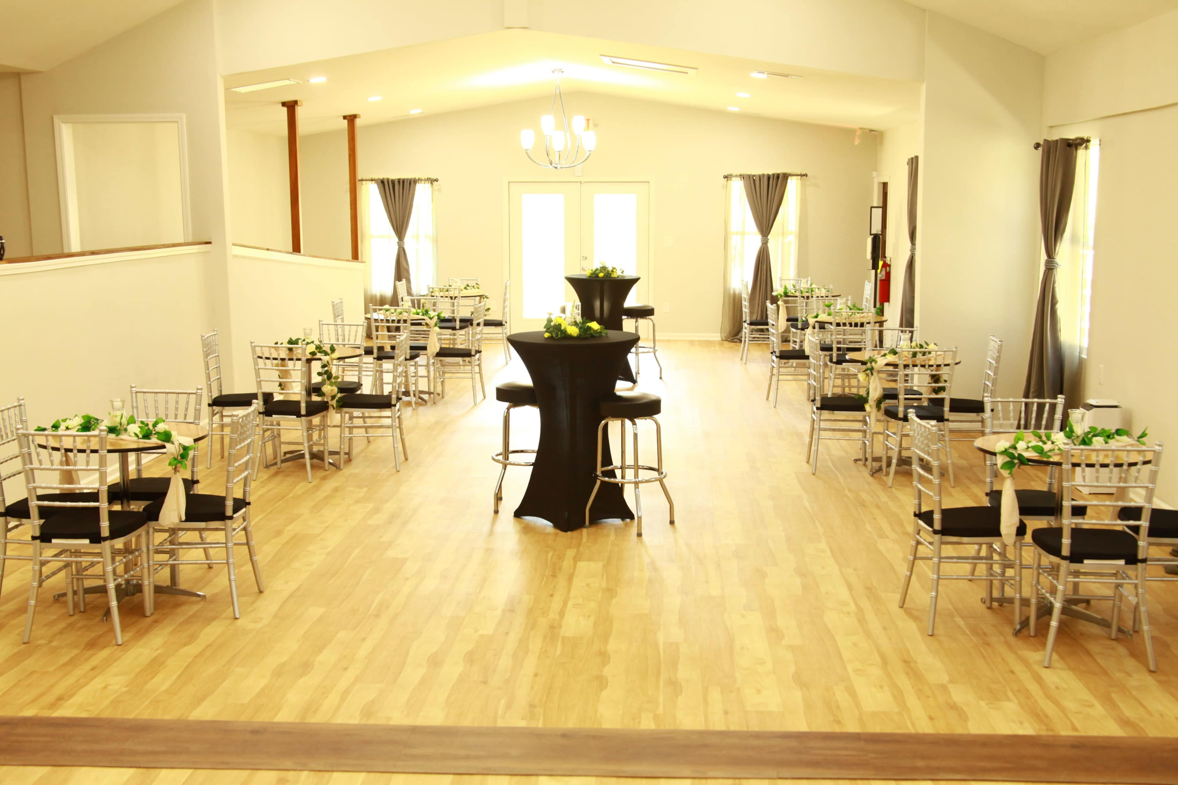 A spacious event hall features several tables with black coverings and floral centerpieces, arranged with chairs and a central standing table.