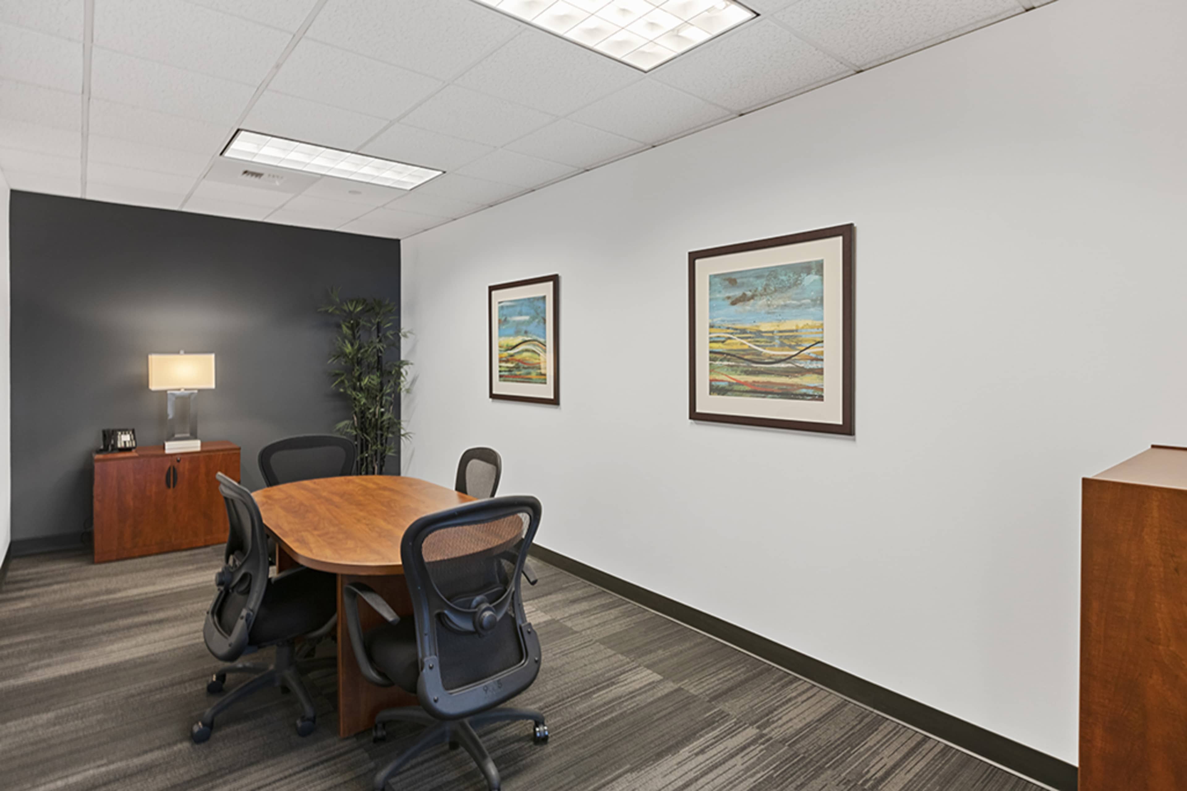 A modern conference room with a round wooden table surrounded by four ergonomic chairs, two framed artworks on the wall, a lamp on a side table, and a tall plant in the corner.