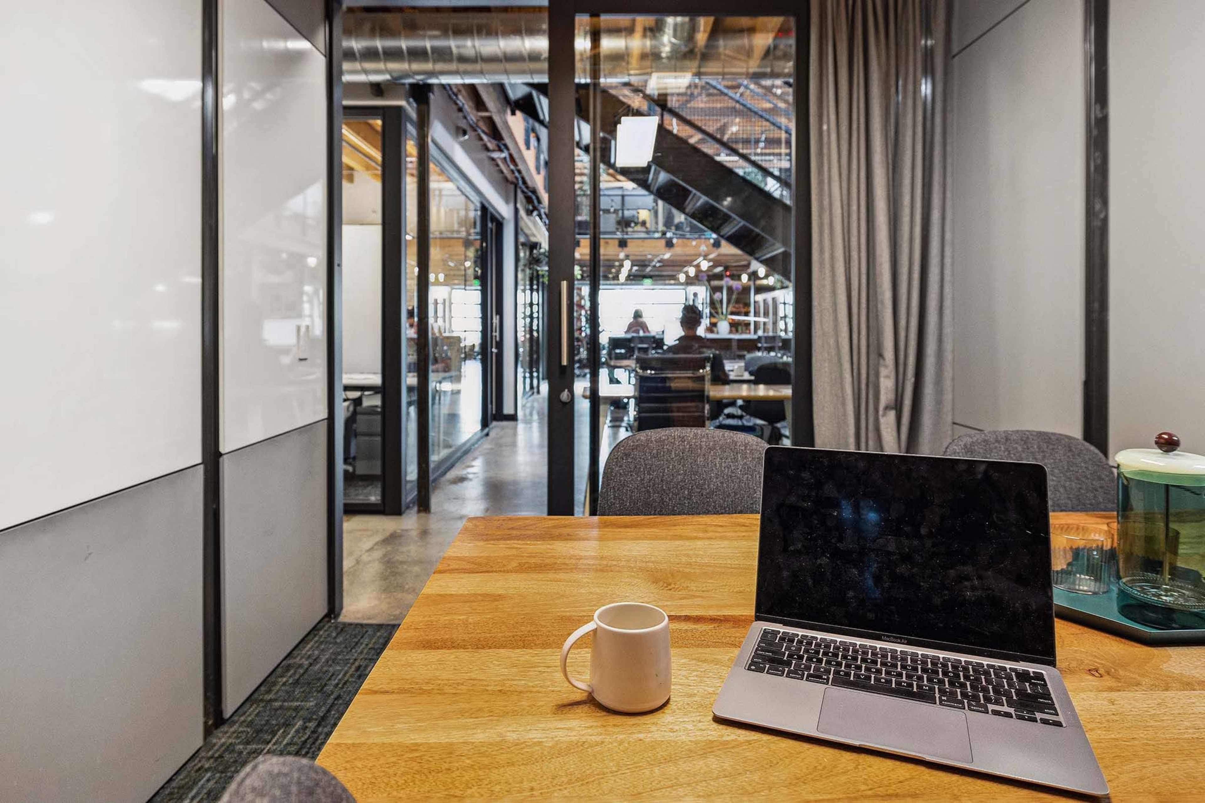A laptop and a white cup are placed on a wooden table in a modern office space with glass walls and a view of a busy workspace.