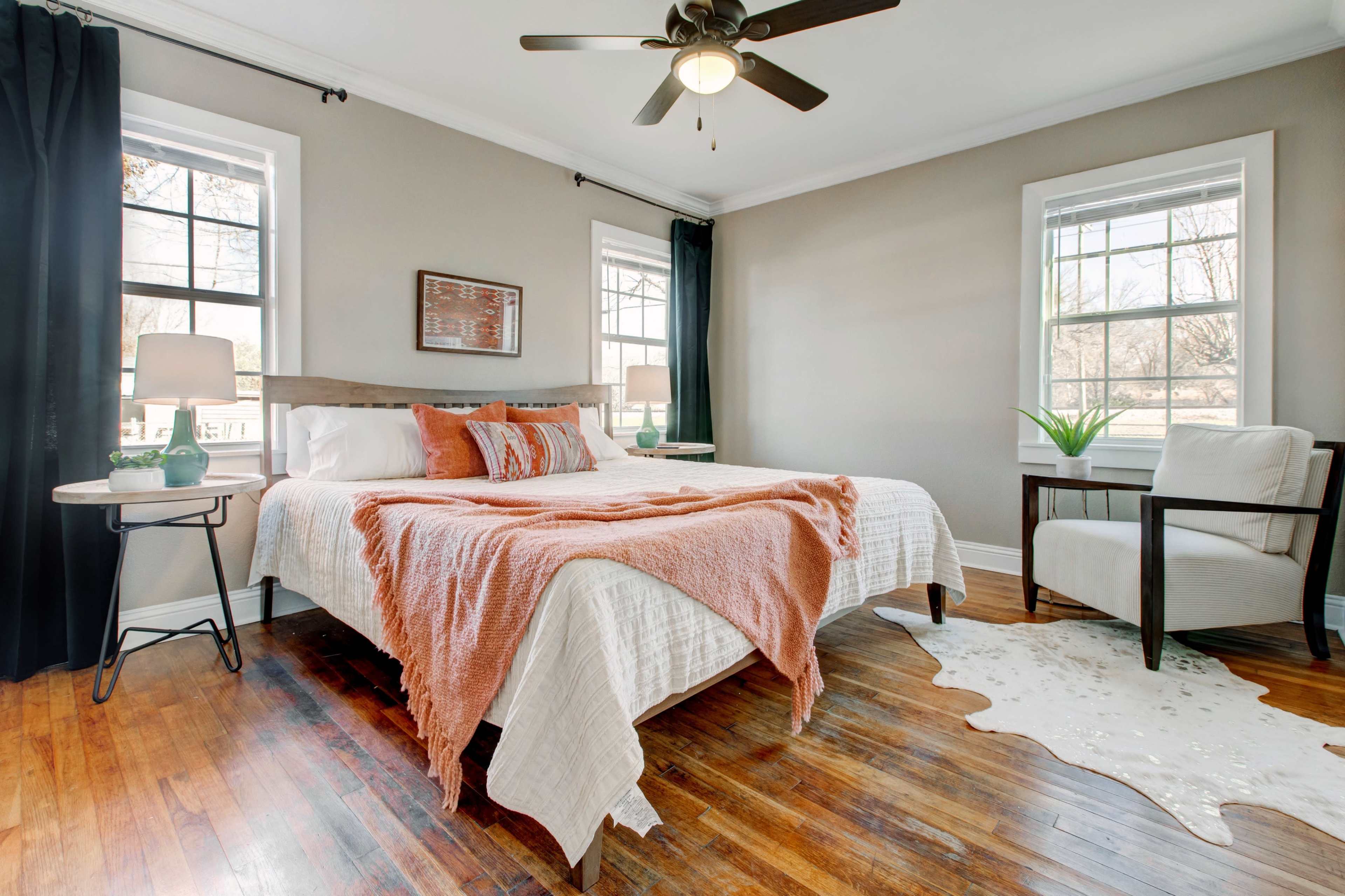 A well-lit bedroom features a bed with an orange throw blanket, a chair, and windows with dark curtains.