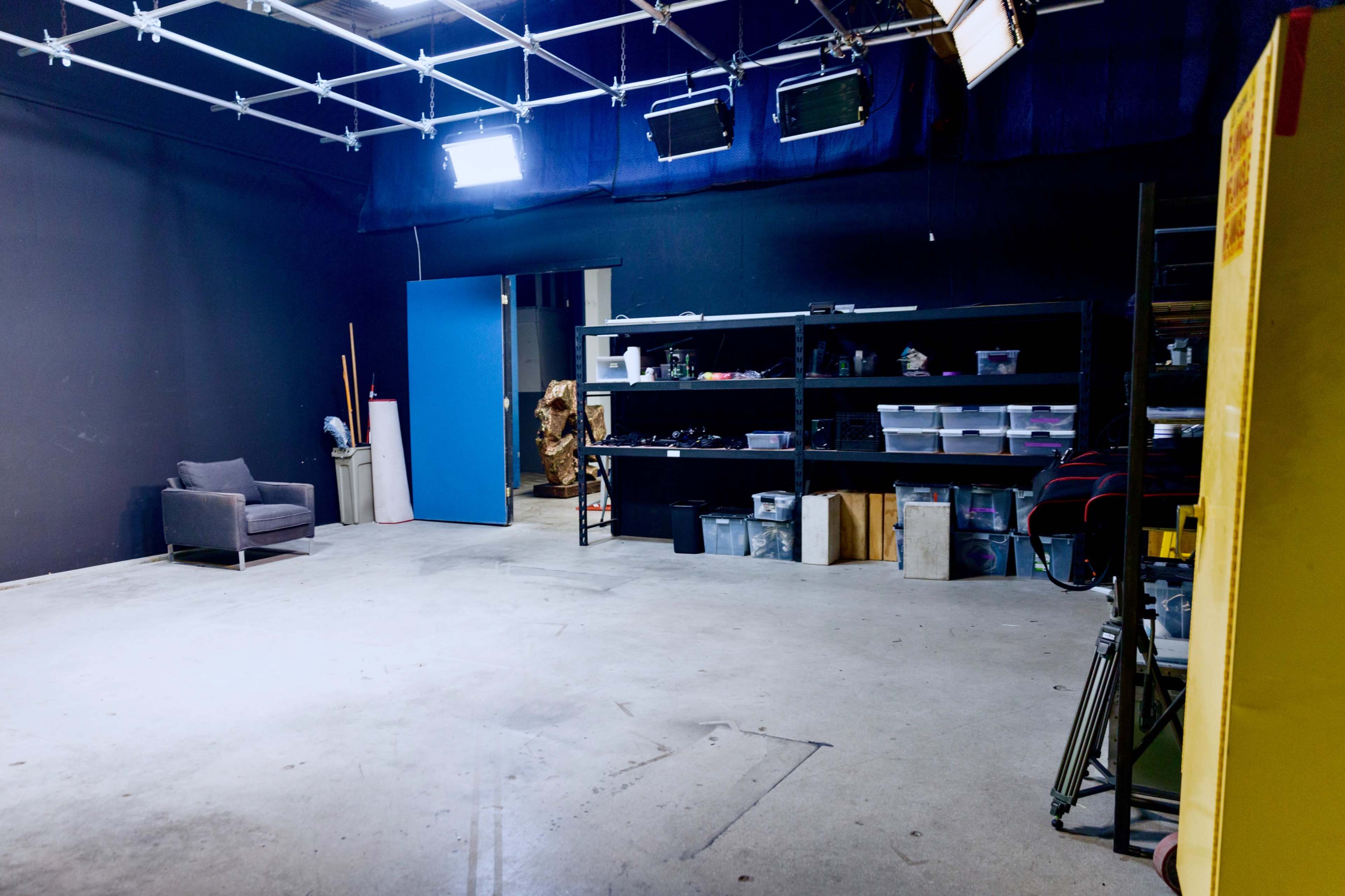 An empty studio space with a gray chair, shelves filled with equipment, and illuminated by overhead lights.