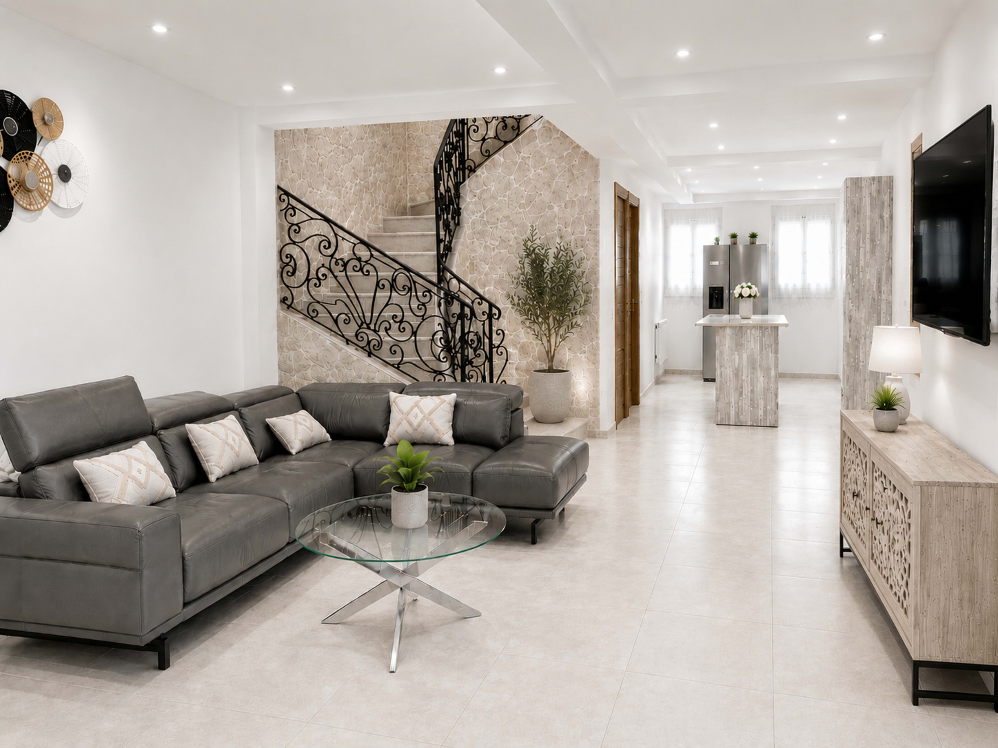 The image shows a modern living space featuring a gray sectional sofa, a round glass coffee table, a decorative wall with metal art, and an open layout leading to a kitchen area with a staircase in the background.