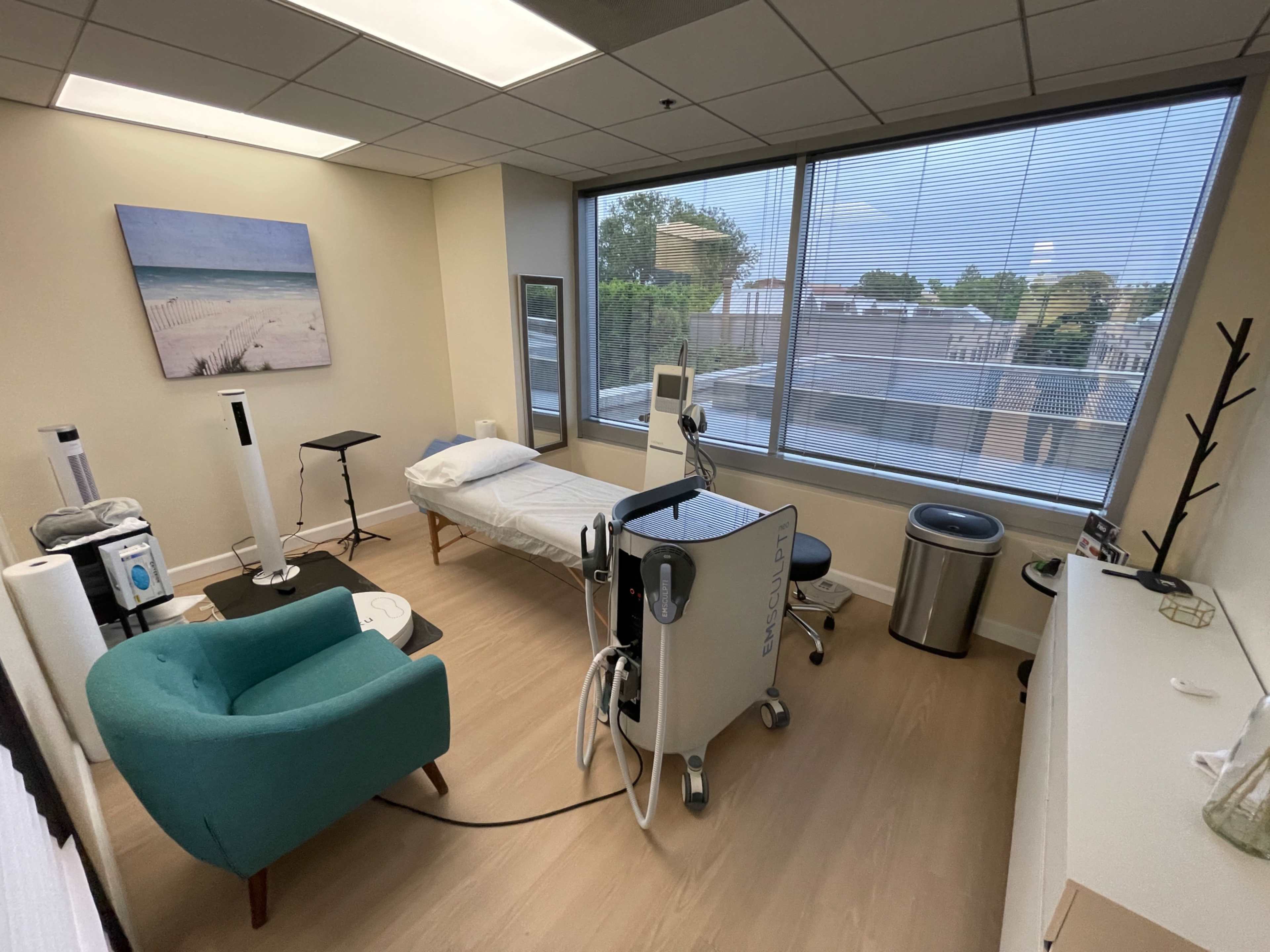 A medical examination room with a bed, chair, and various medical equipment near a large window.