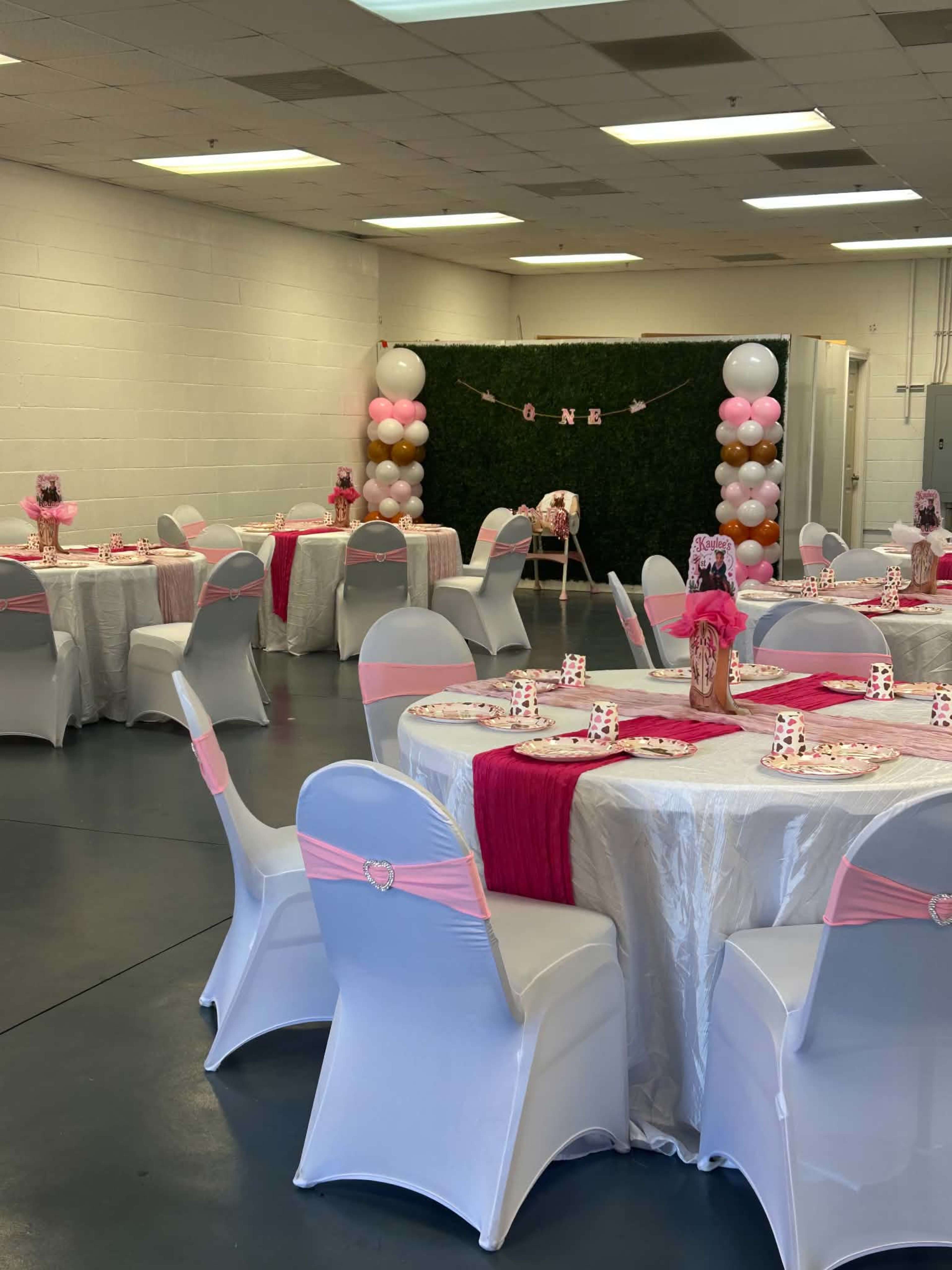 The image shows a decorated event space with round tables set for a celebration, featuring white tablecloths, pink accents, and a backdrop of balloons and greenery.