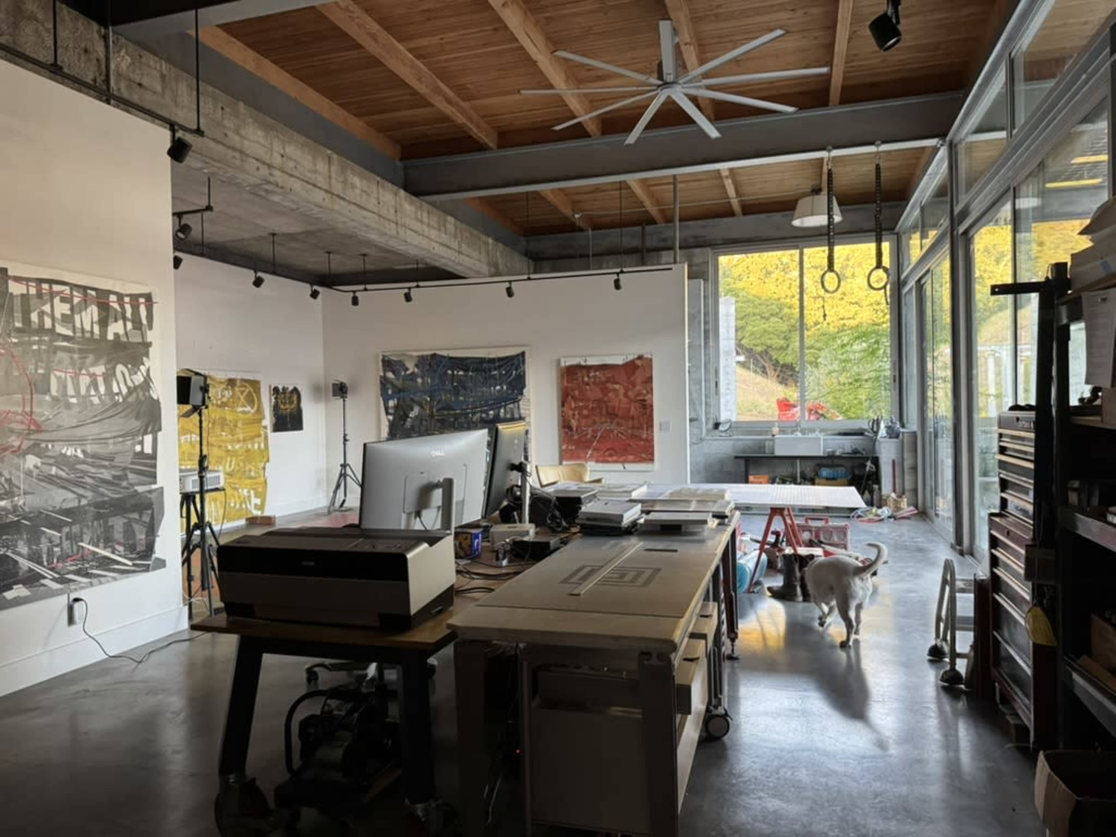 The image shows a spacious modern workspace with large windows, a central workstation, and a cat walking through the room.
