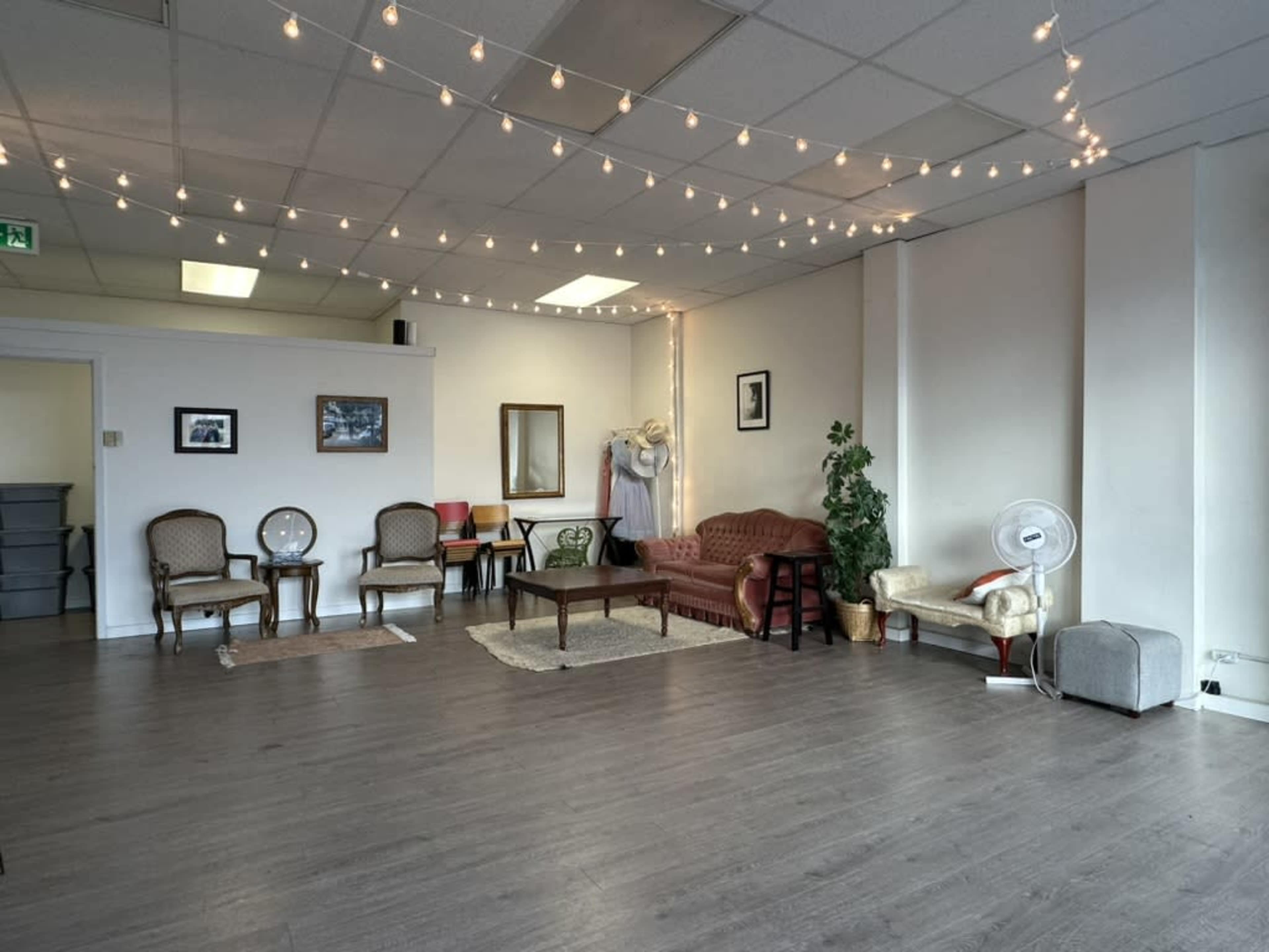 A spacious room with wooden flooring, decorated with string lights, featuring several seating areas, a table, and a mirror.