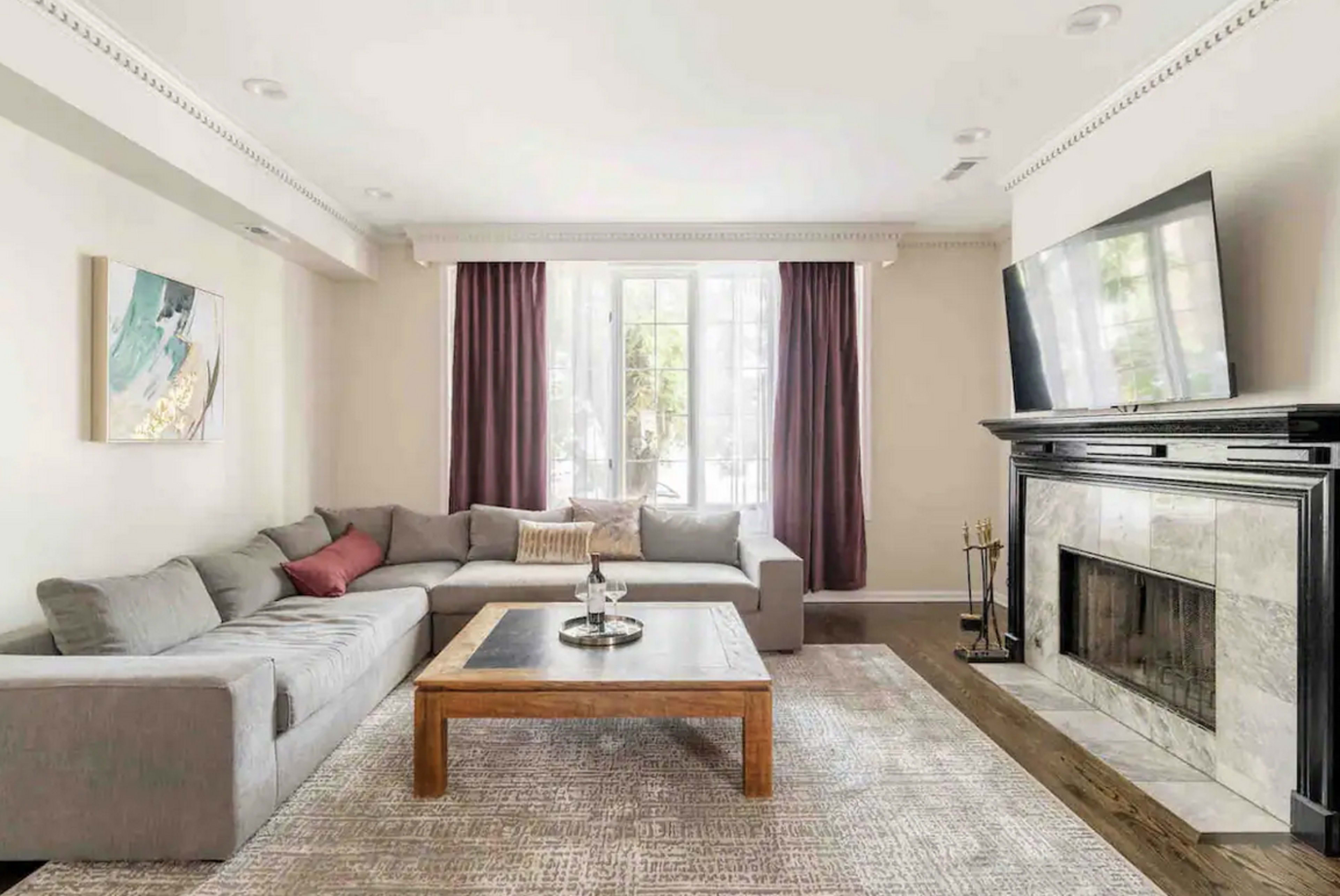 A modern living room features a gray sectional sofa, a wooden coffee table, and a marble fireplace, with large windows dressed in light curtains.