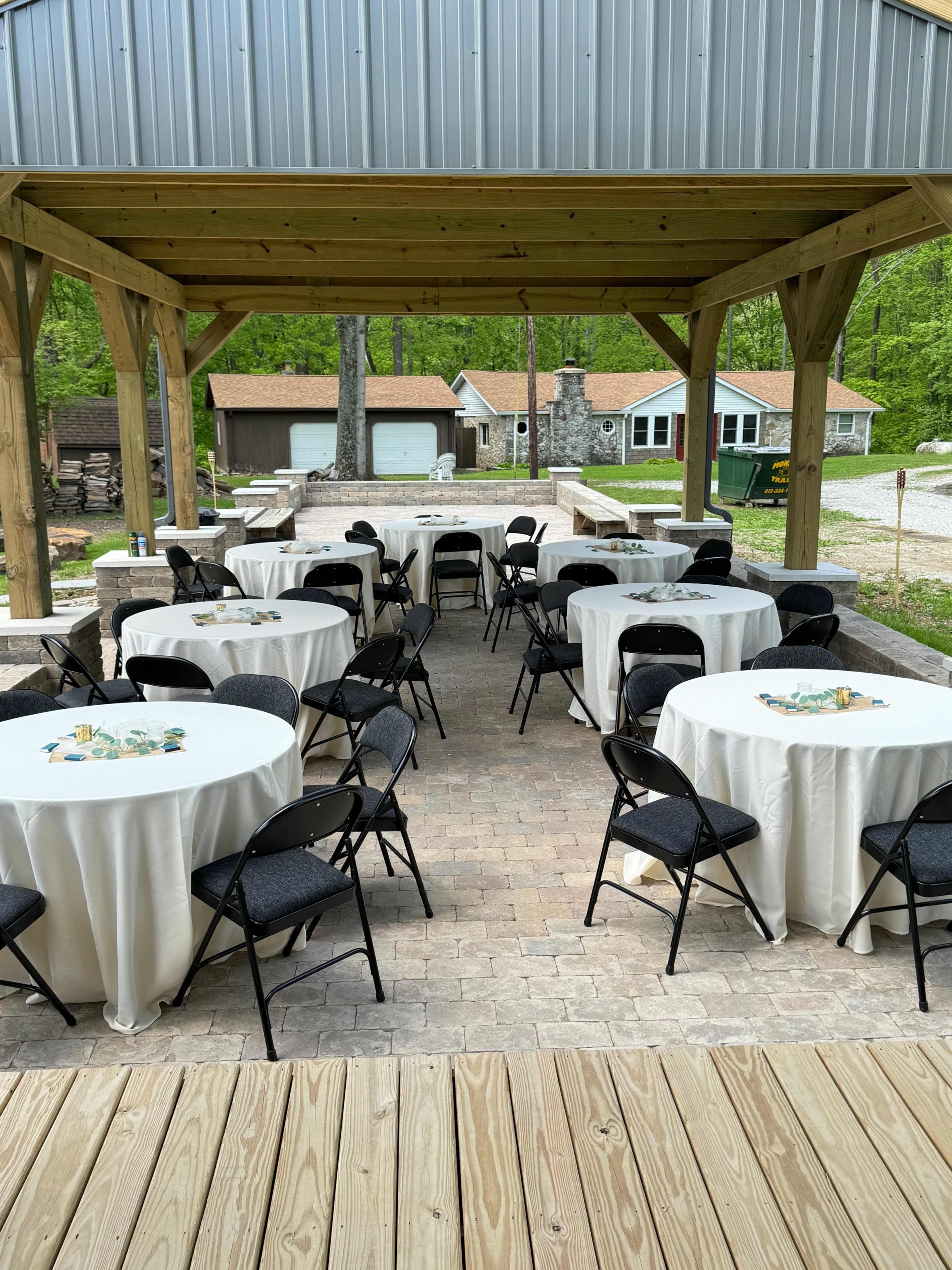 A covered outdoor area with several round tables set with white tablecloths and black chairs, surrounded by greenery and a stone fire pit in the background.