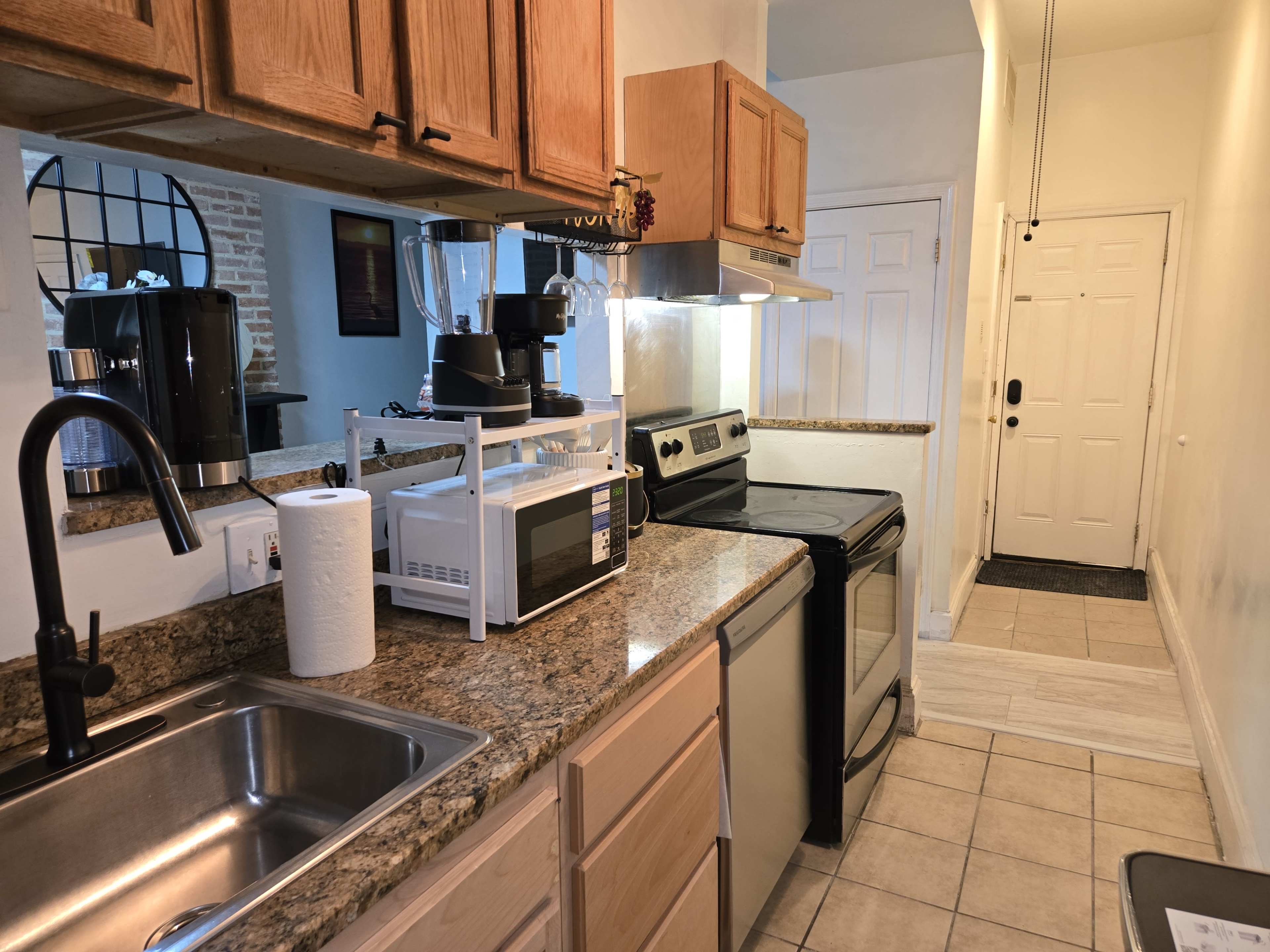 The image shows a kitchen with wooden cabinets, a stainless steel stove, a microwave, and a sink, alongside a blender and a coffee maker on the countertop.