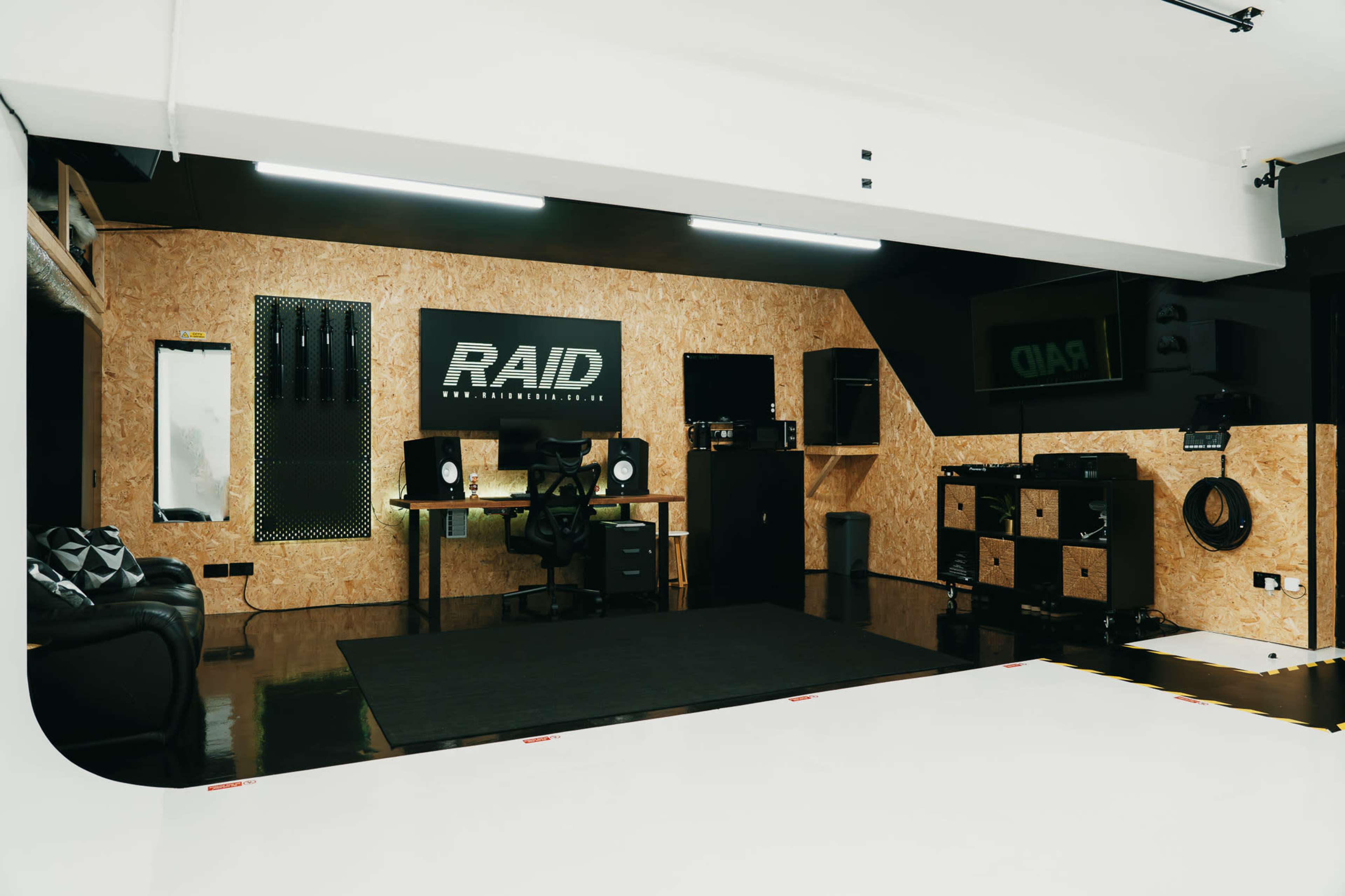 The image shows a modern studio space with a black and orange color scheme, featuring a desk setup with computer equipment, storage units, and seating.