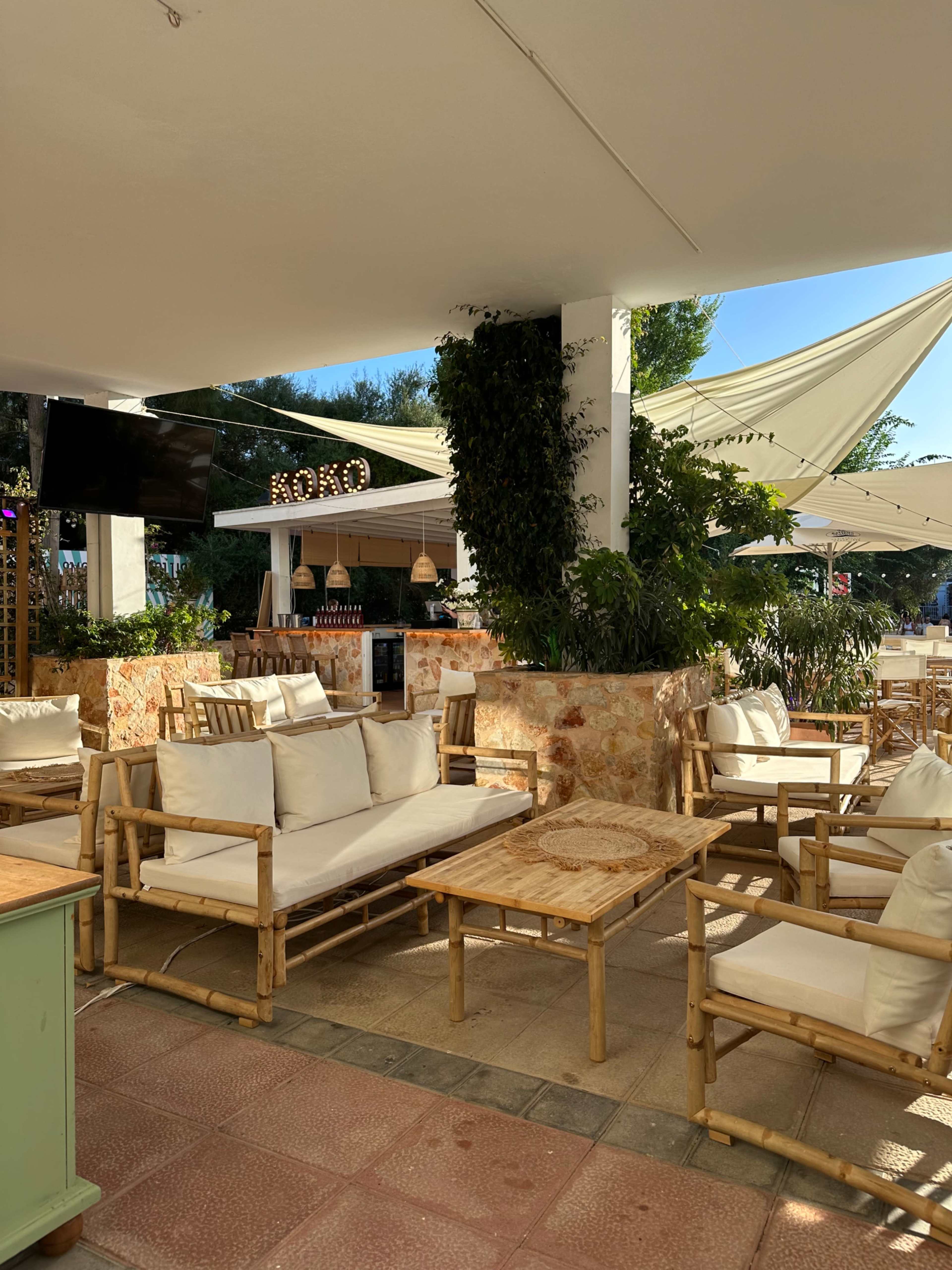 The image shows a modern outdoor lounge area with bamboo furniture, soft cushions, and shaded seating beneath a canopy.