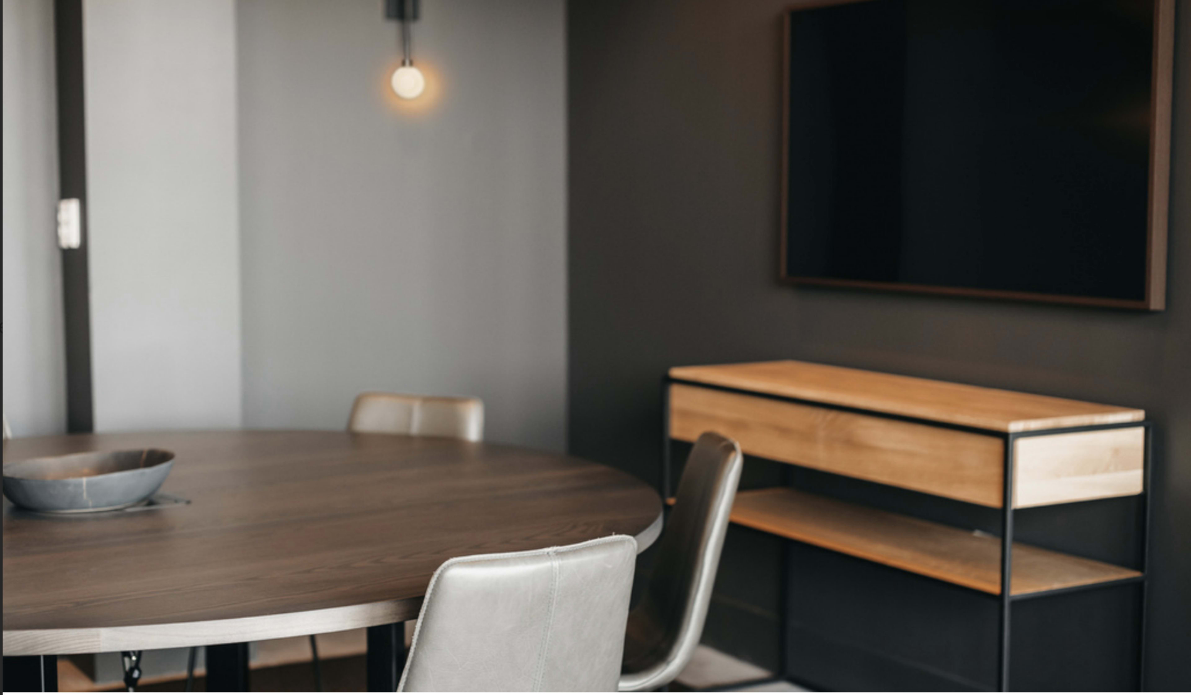 The image shows a modern dining area featuring a round wooden table, four metal and upholstered chairs, and a wooden sideboard next to a wall-mounted television.