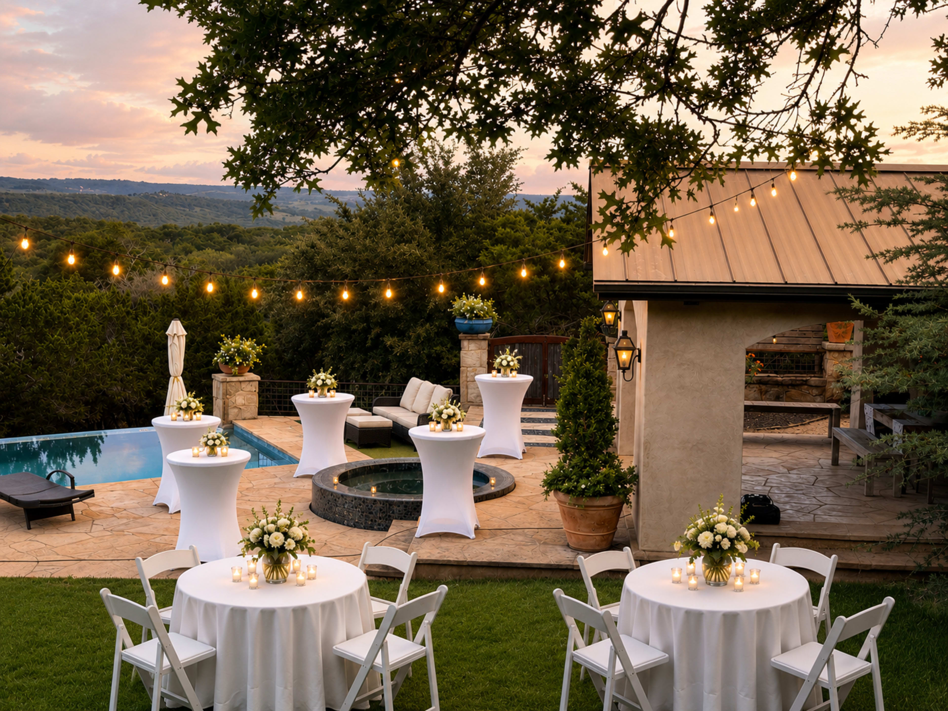 A beautifully arranged outdoor event space features white tables with floral centerpieces, elegant cocktail tables by a pool, and string lights illuminating the scene under a sunset sky.