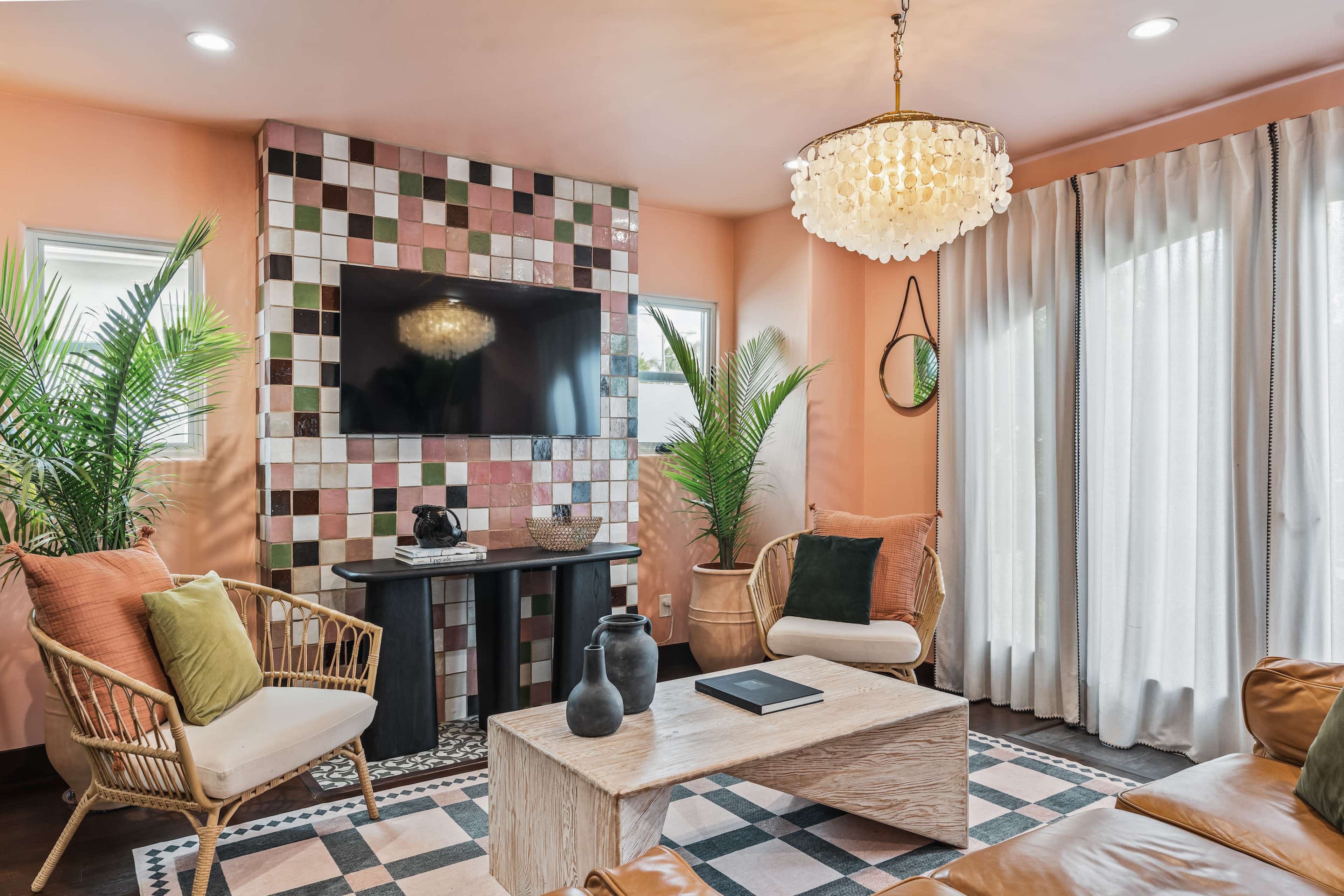The room features a combination of seating, a large television mounted on a colorful tiled wall, and plants arranged around a wooden coffee table.