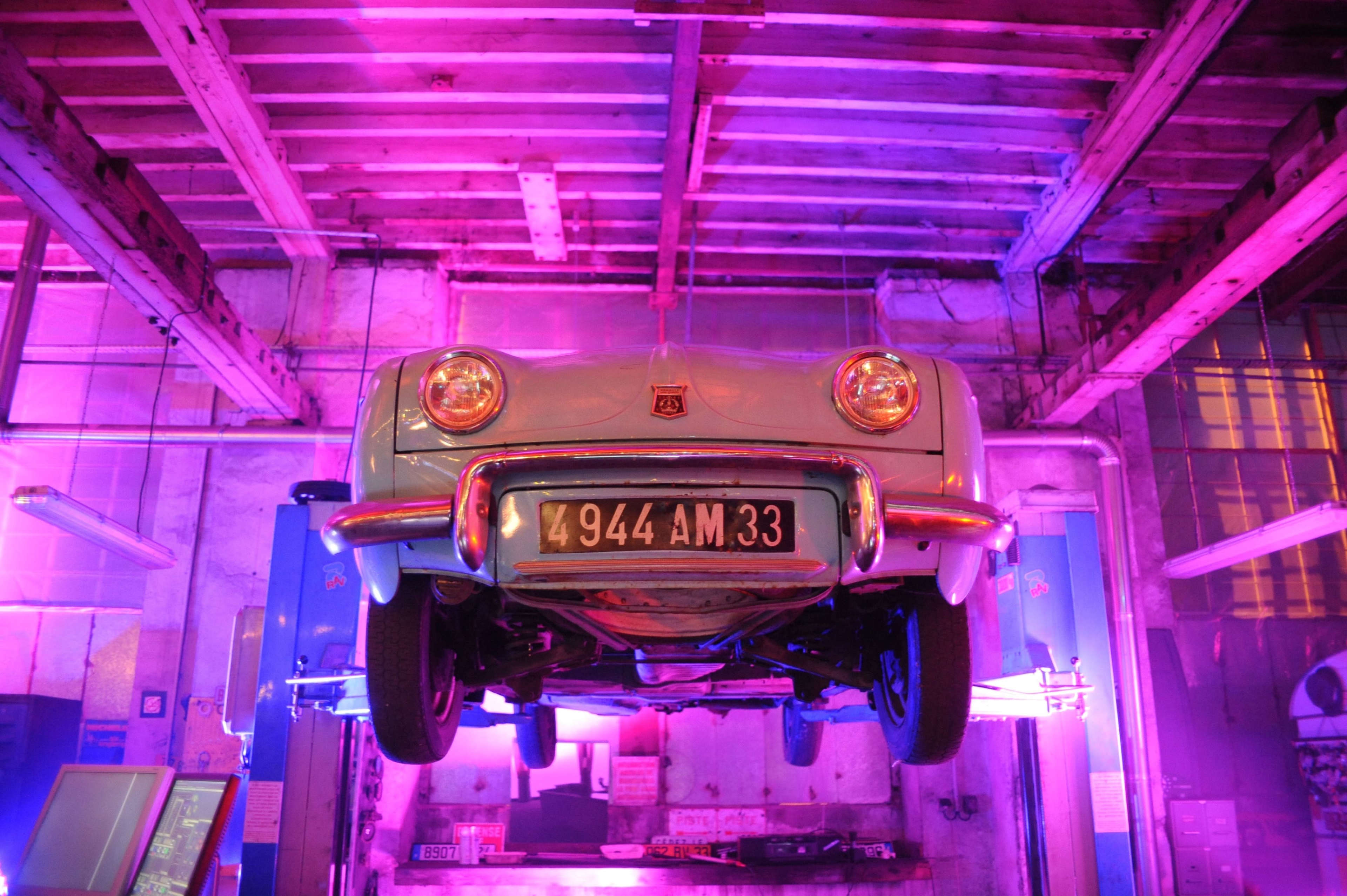 A vintage car is suspended upside down in a brightly lit garage with purple lighting.
