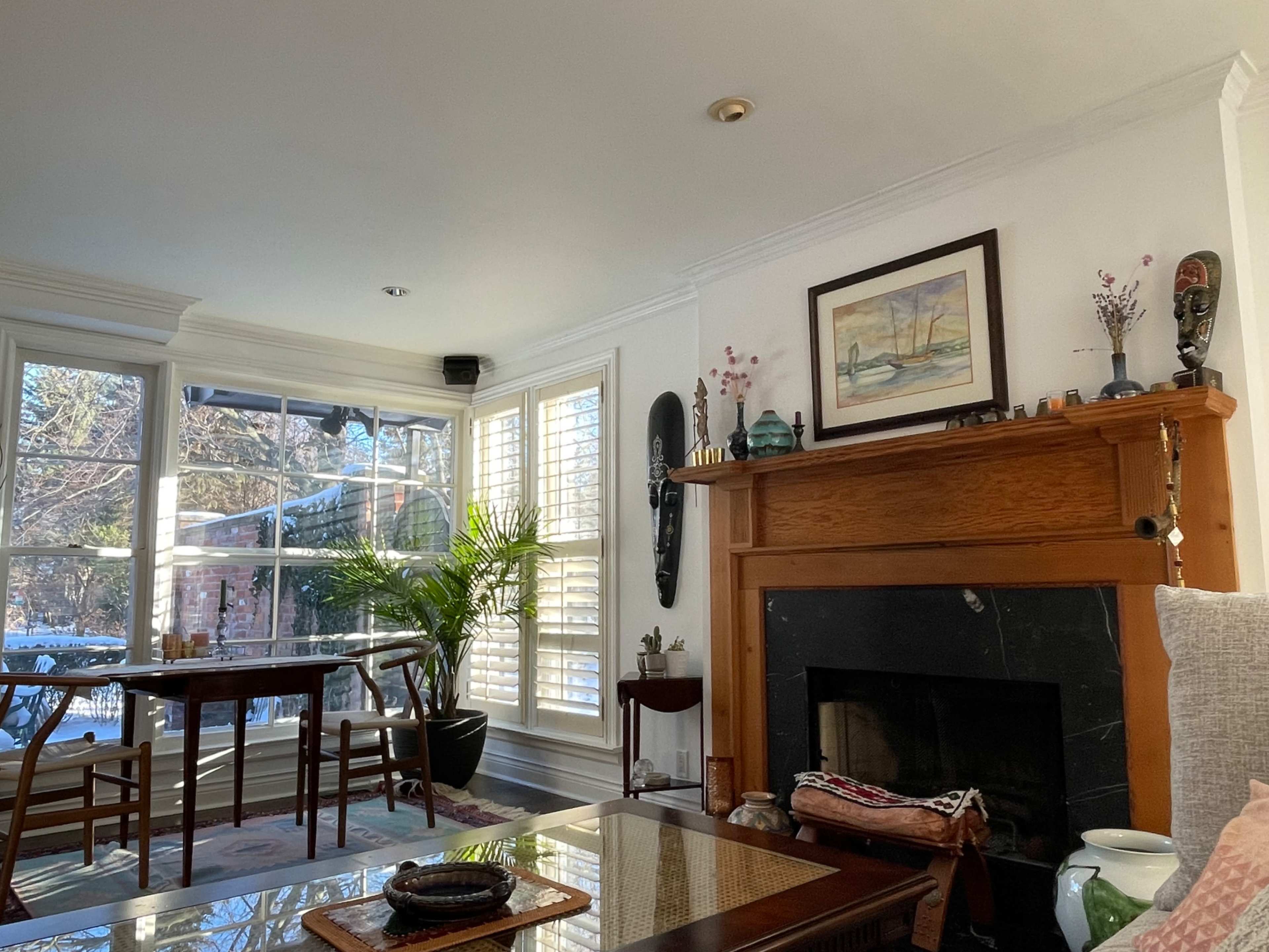 A sunlit living room features a wooden fireplace, a dining table, and large windows with plantation shutters overlooking a snowy outdoors.