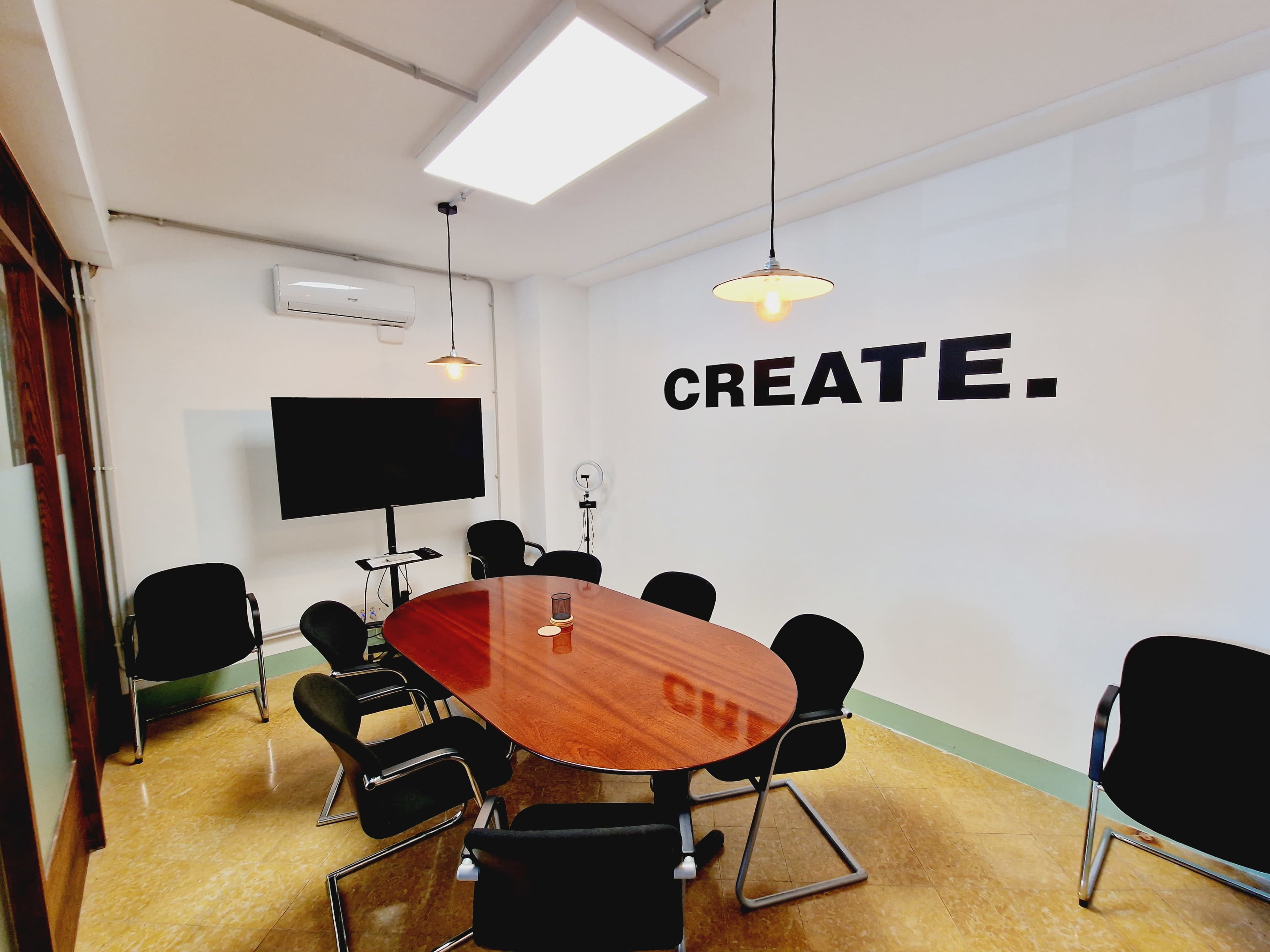 A conference room features a large wooden table surrounded by black chairs, with a wall displaying the word "CREATE" and a screen mounted nearby.