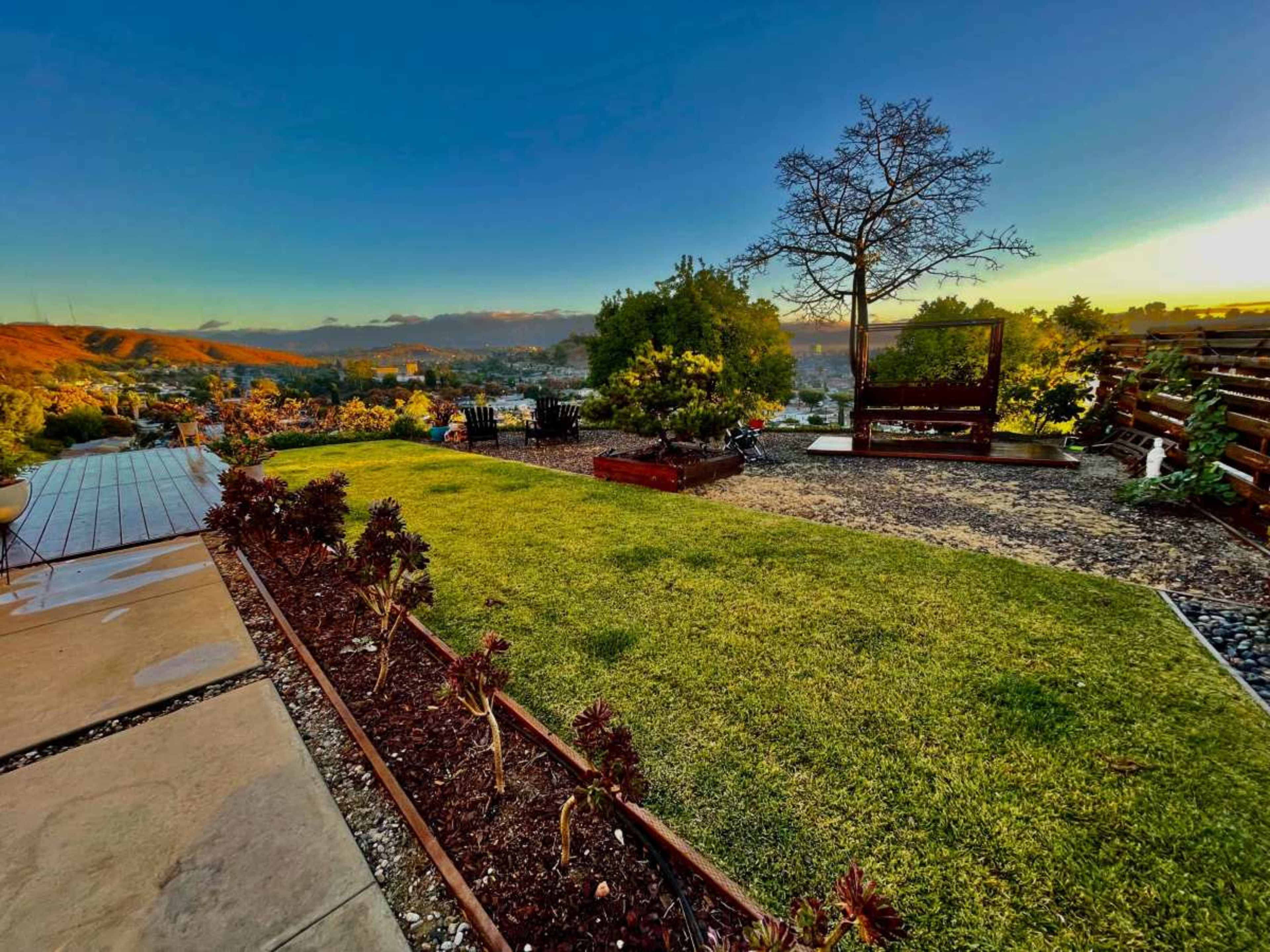 The image shows a landscaped garden with green grass, bordered by flower beds, overlooking a city view at sunset.
