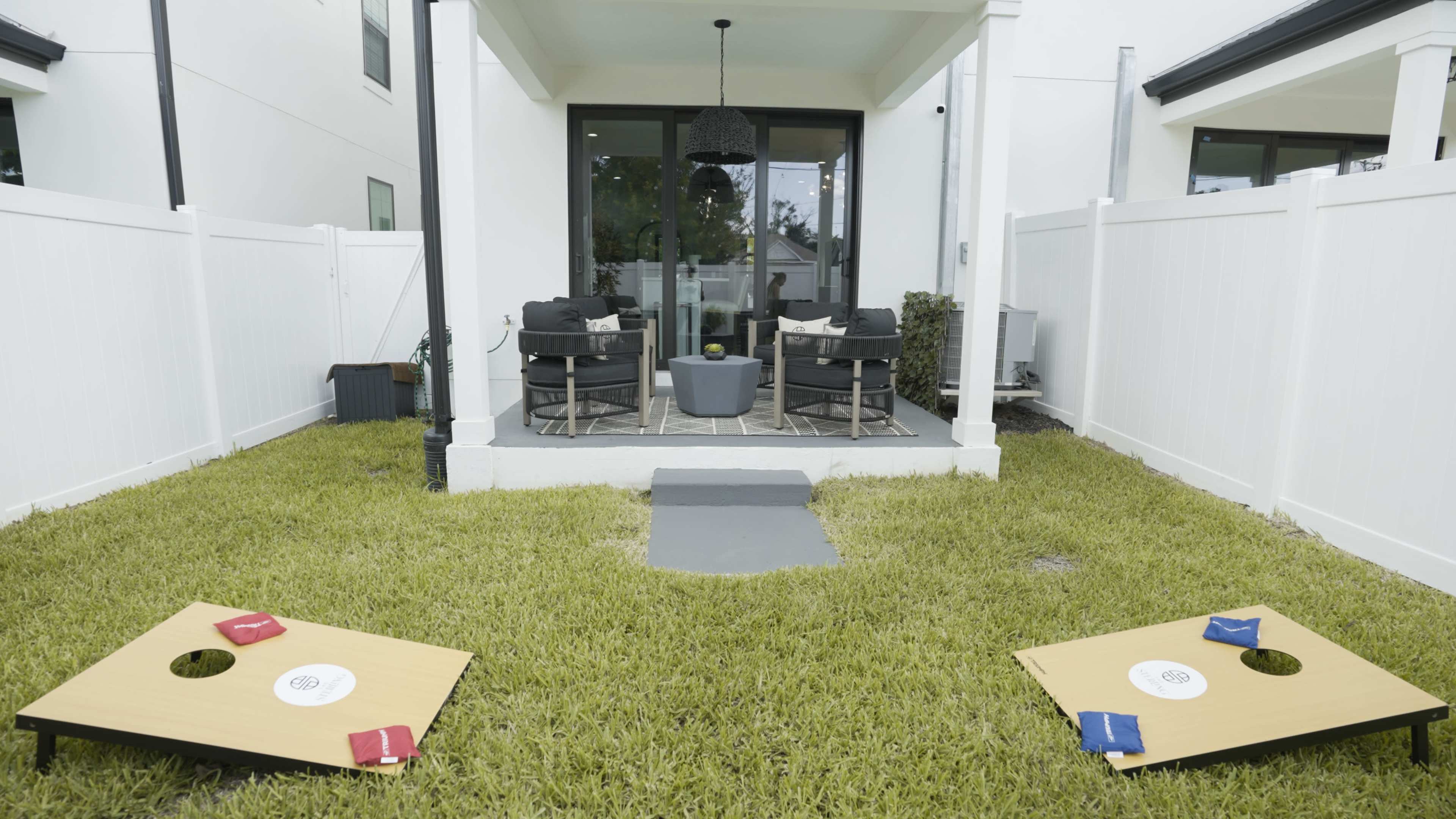 The image shows a backyard with two cornhole boards and bean bags set up on a grassy area in front of a patio with seating.