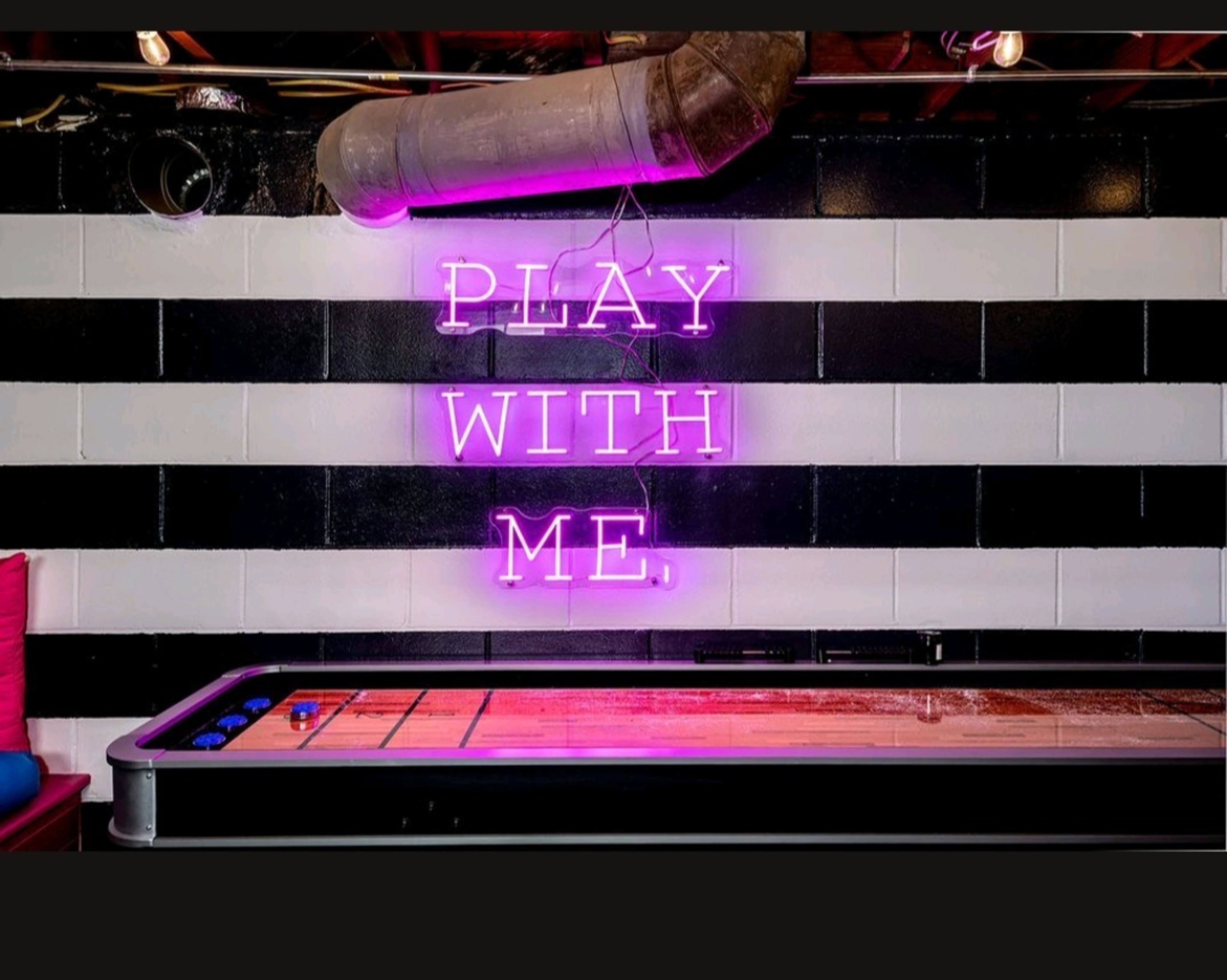 A pink neon sign that reads "PLAY WITH ME," is displayed on a black and white striped wall above a shuffleboard table.