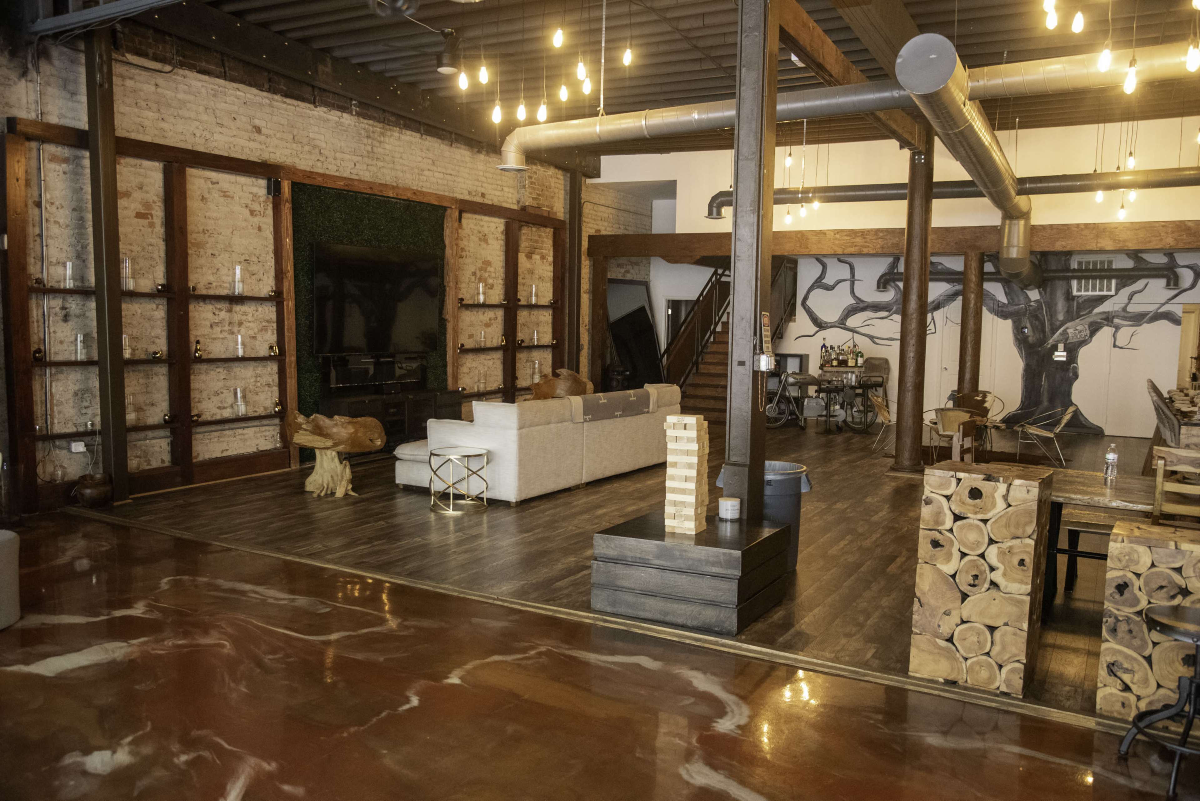 The image shows a spacious, modern interior with a mix of wooden and industrial elements, featuring a living area with a sofa, decorative wooden accents, and a unique floor design.