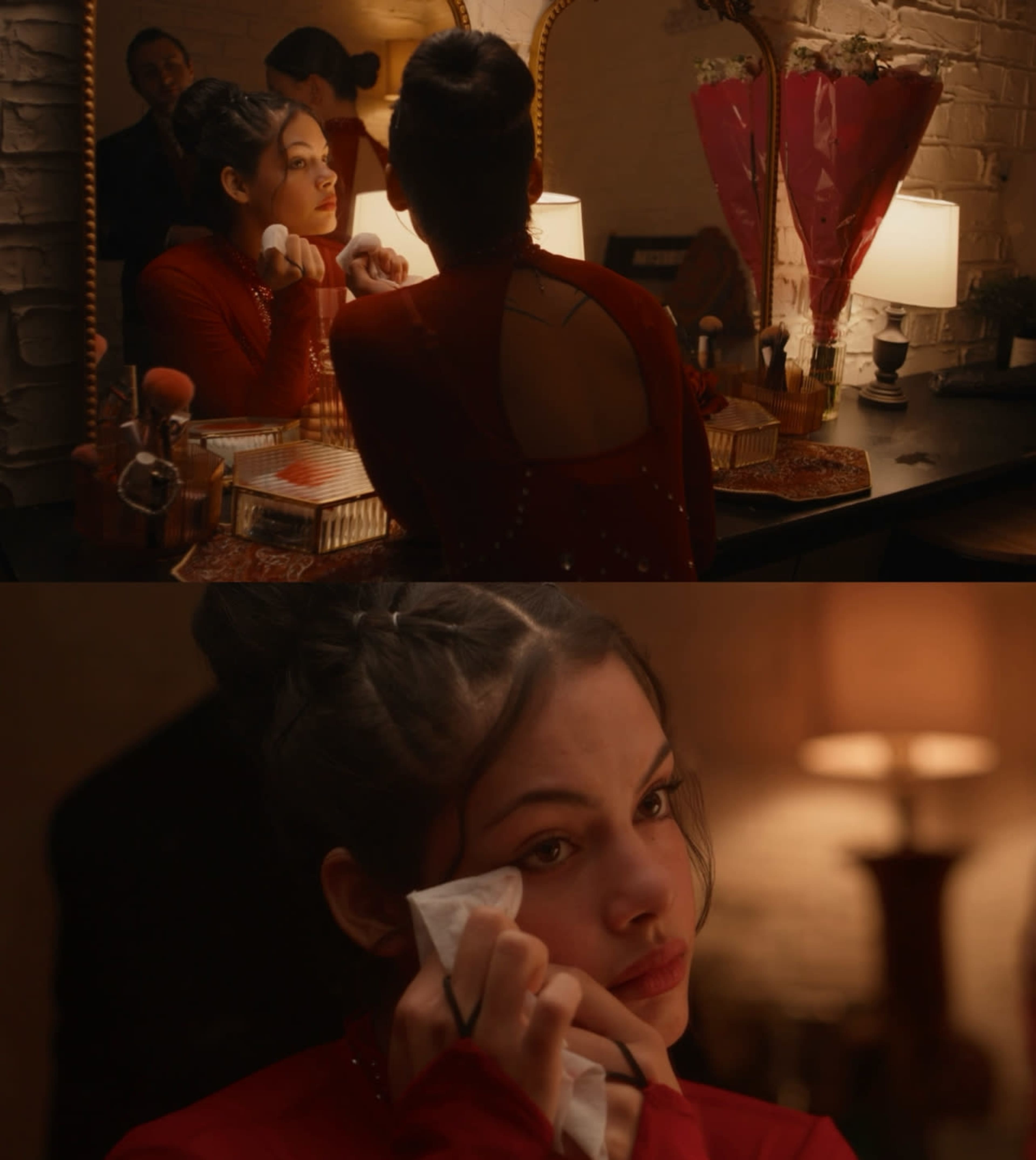 A woman in a red dress is seen at a vanity, removing makeup in front of a mirror with soft lighting.