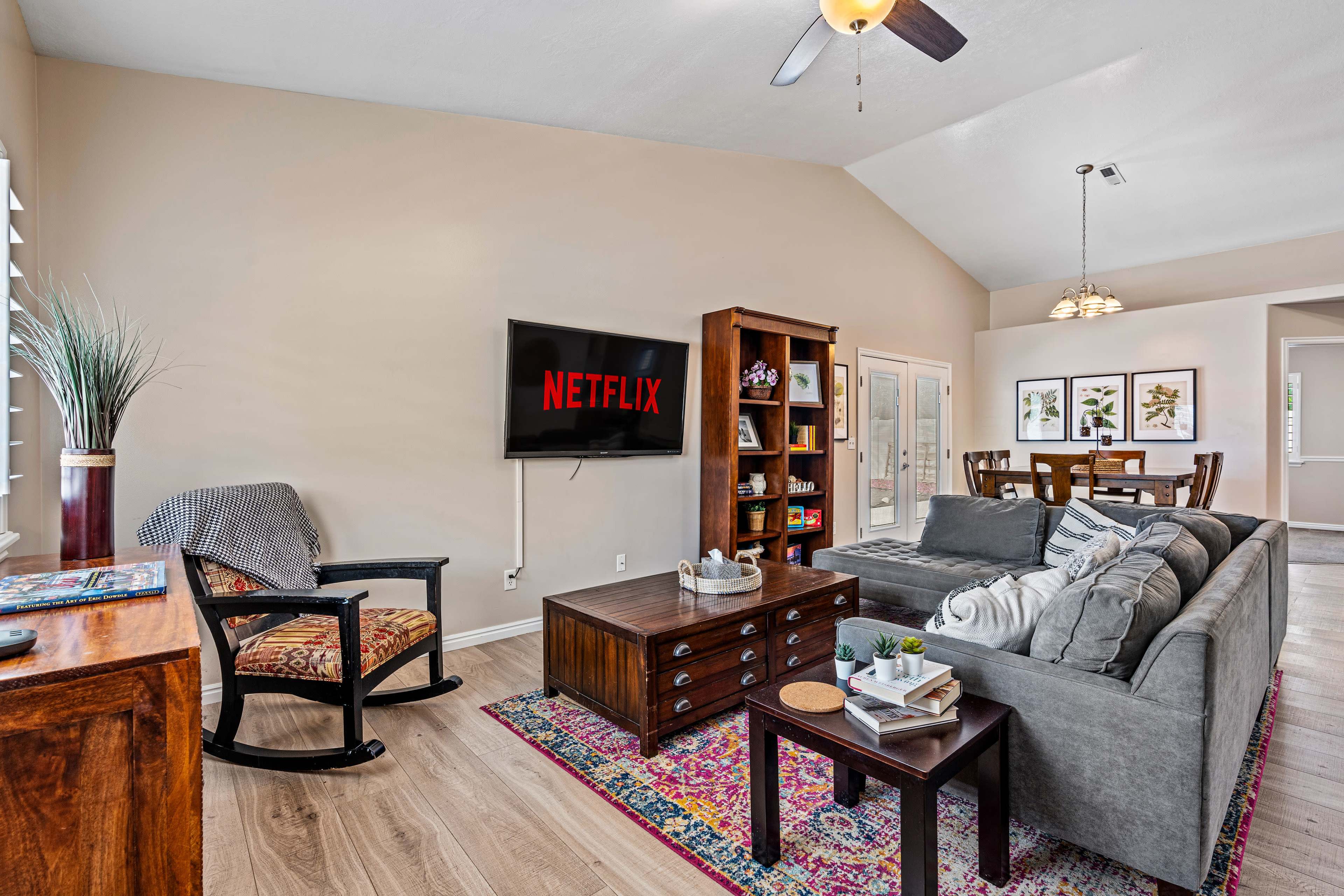 The living room features a comfortable seating area with a gray couch, a wooden coffee table, and a television displaying the Netflix logo, along with a dining area visible in the background.